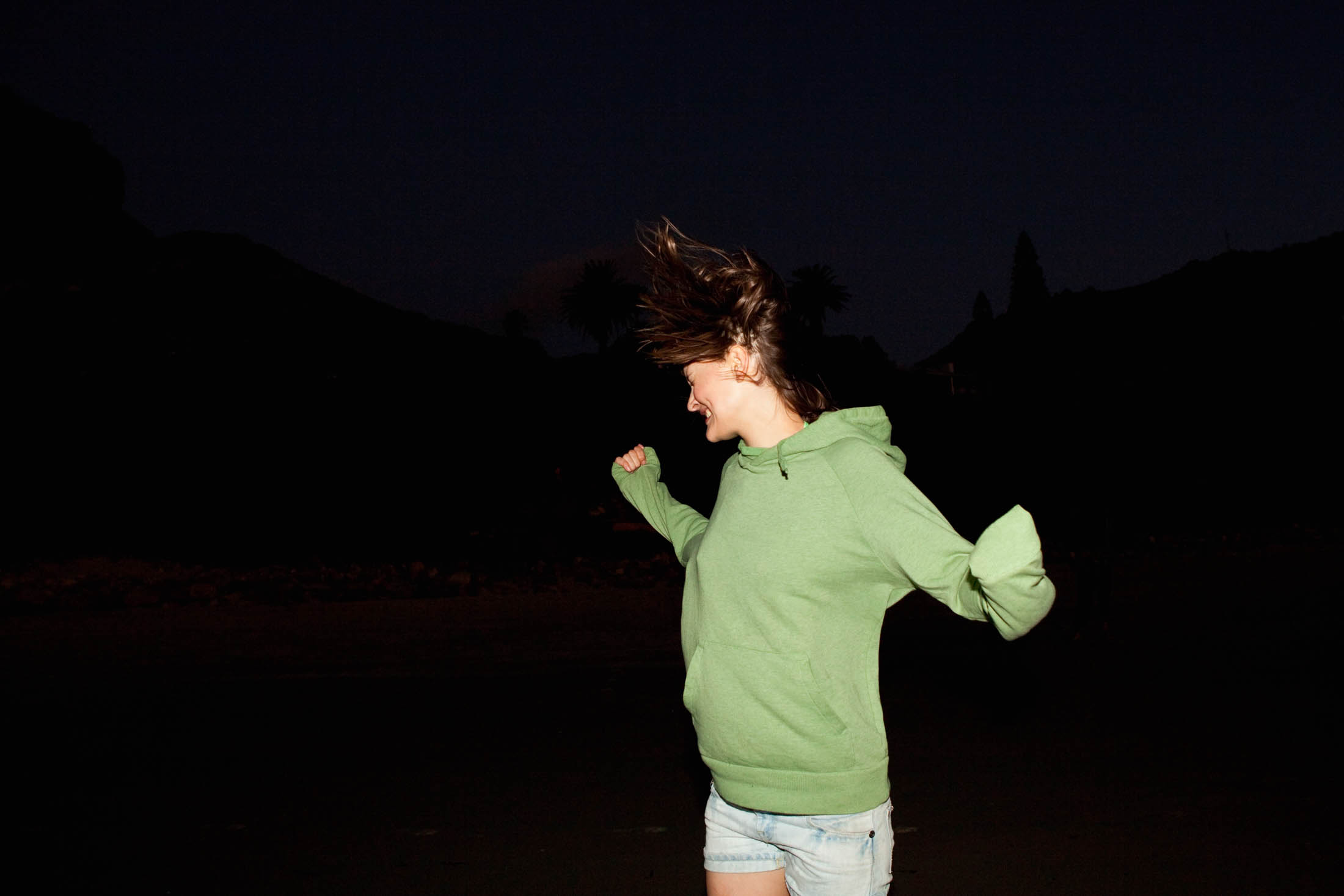 Eine junge Frau in einem grünen Kapuzenpullover und Jeansshorts lacht fröhlich unter einem schwach beleuchteten Abendhimmel, während ihr Haar wild im Wind weht. © Fotografie Tomas Rodriguez