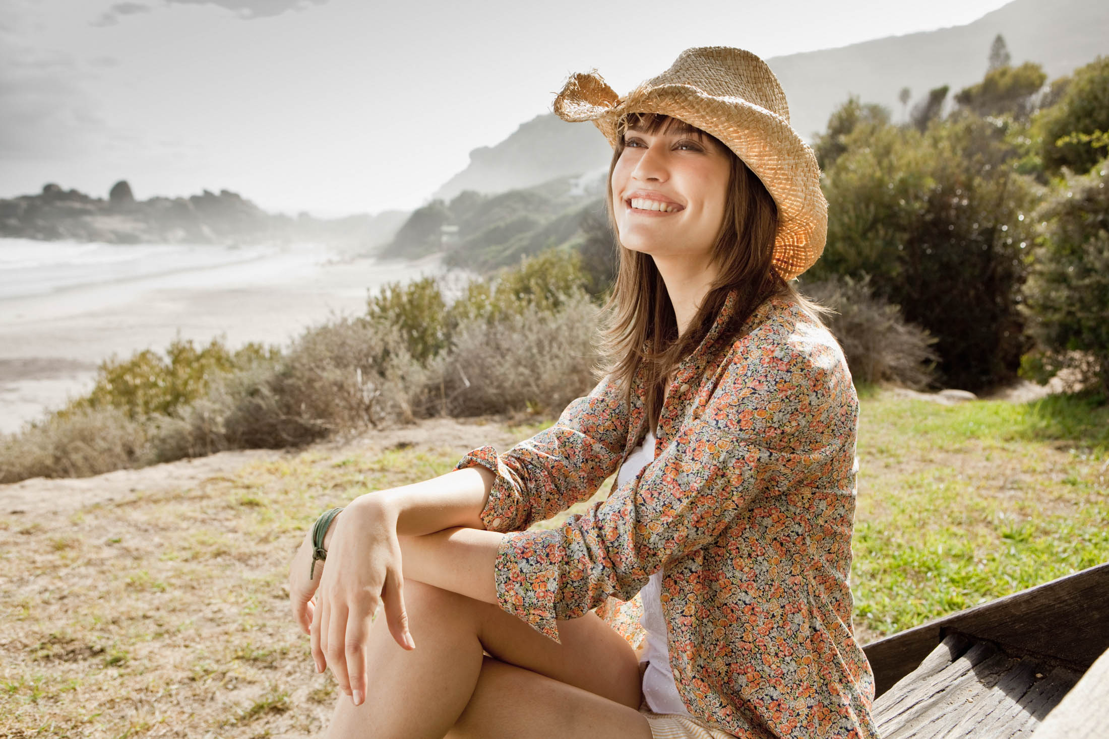 Eine glückliche junge Frau mit Strohhut und geblümter Jacke sitzt auf einer Holzbank an einem sonnigen Strand und lächelt in die Kamera. © Fotografie Tomas Rodriguez