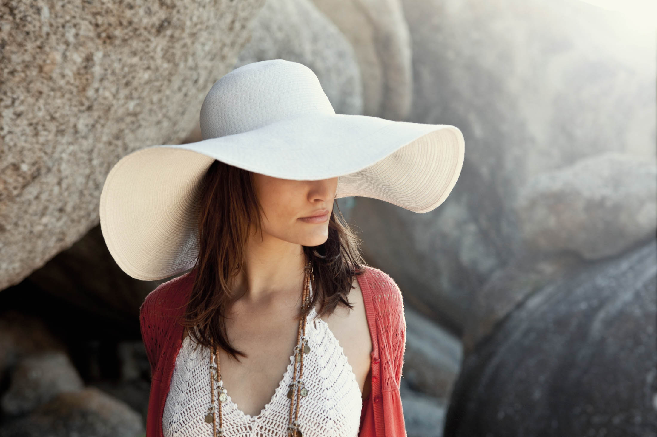 Eine Frau mit einem breitkrempigen weißen Hut und einem roten Oberteil steht zwischen großen Felsen, schützt ihr Gesicht teilweise mit ihrem Hut und blickt nach unten. © Fotografie Tomas Rodriguez