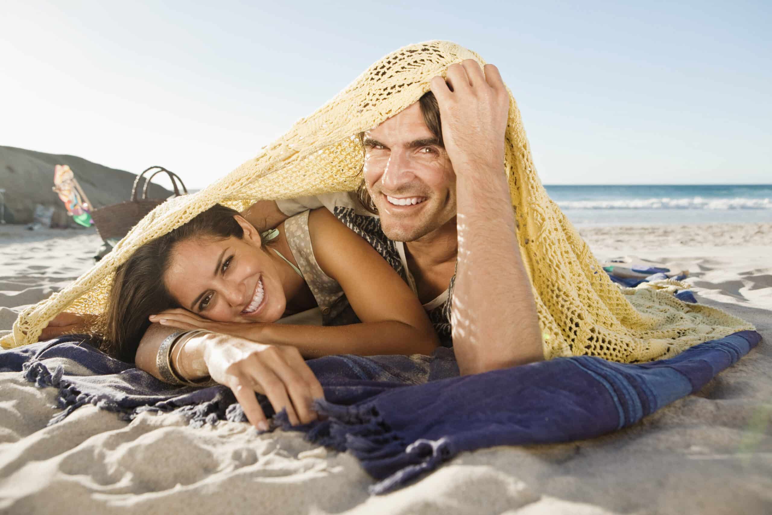 Ein lächelndes Paar liegt am Strand unter einer gelben Strickdecke. Die Frau ruht mit dem Kopf auf ihren Armen und der Mann hält den Rand der Decke hoch. Sie liegen auf einem gestreiften Strandtuch mit dem Meer und einem klaren Himmel im Hintergrund. © Fotografie Tomas Rodriguez