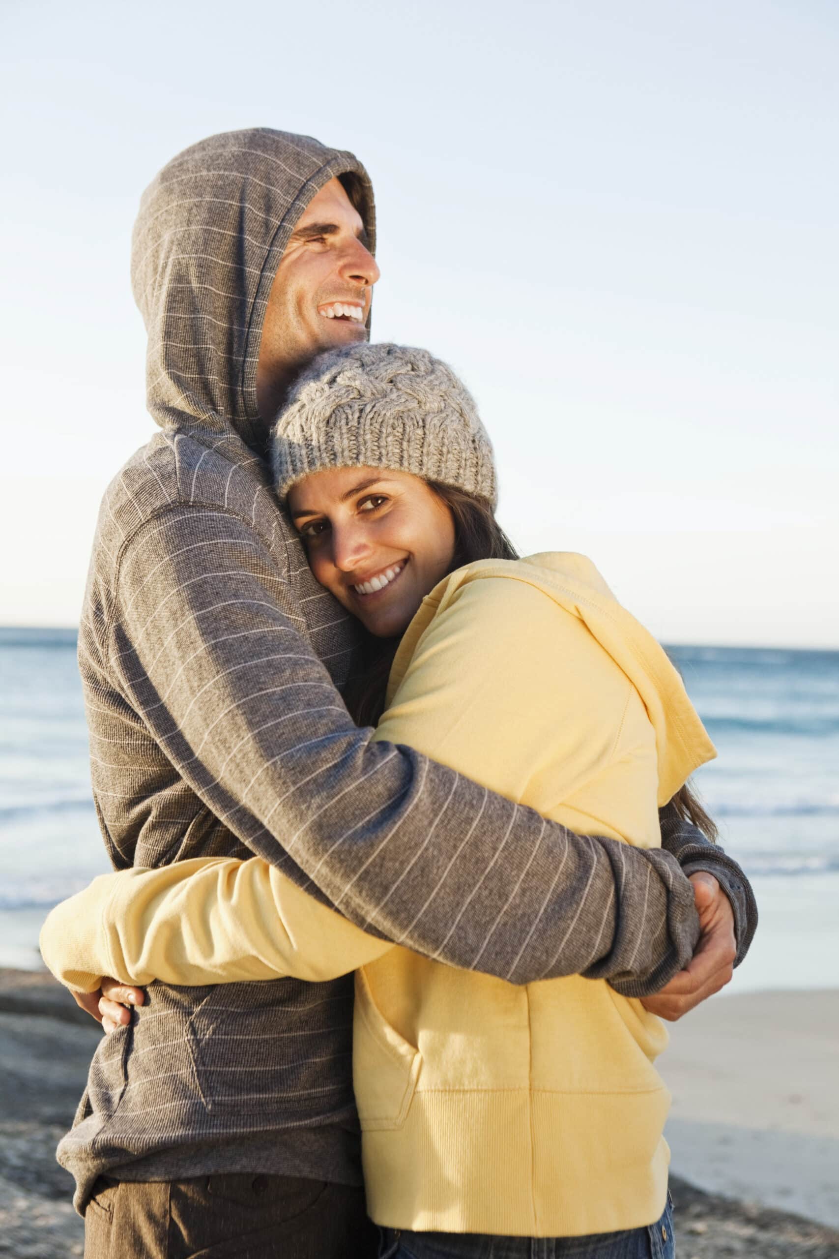 Ein warm gekleidetes Paar umarmt sich am Strand. Der Mann trägt einen grau gestreiften Kapuzenpullover und die Frau einen gelben Kapuzenpullover mit einer grauen Mütze. Beide lächeln, im Hintergrund sind Strand und Meer zu sehen. © Fotografie Tomas Rodriguez