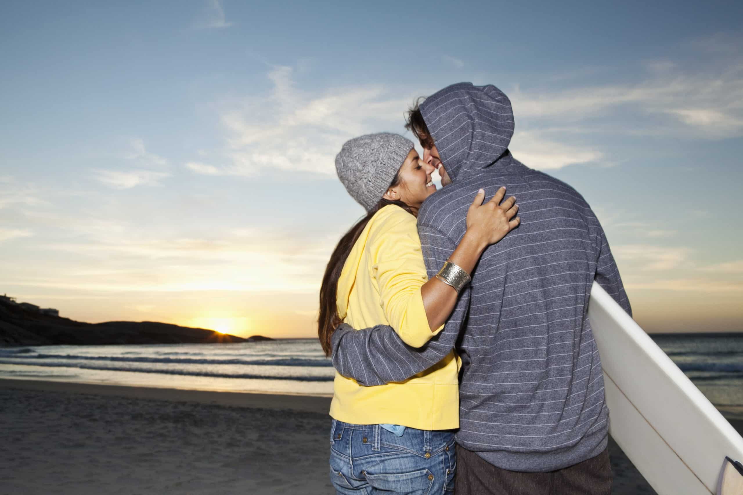 Ein Paar umarmt sich bei Sonnenuntergang an einem Strand. Die Frau trägt eine gelbe Jacke und eine graue Mütze, während der Mann einen gestreiften Kapuzenpullover trägt und ein Surfbrett hält. Der Himmel ist klar und im Hintergrund geht die Sonne über dem Meer unter. © Fotografie Tomas Rodriguez