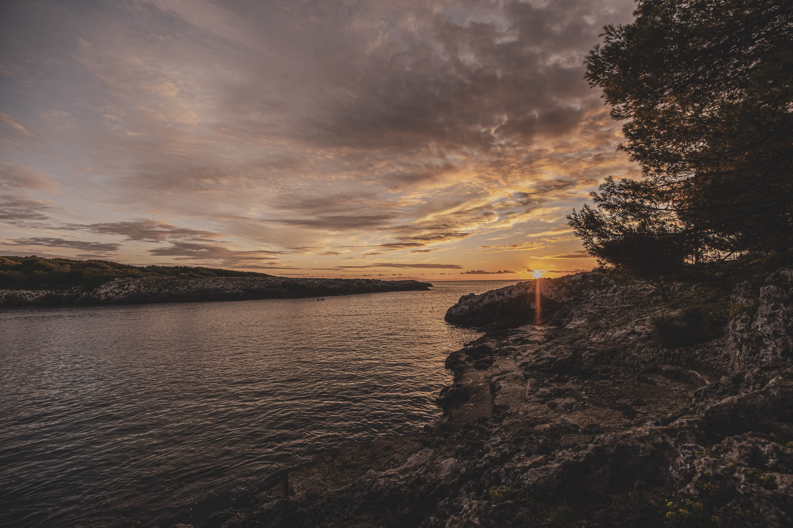 Sonnenuntergang über einem ruhigen Meer, betrachtet von einer felsigen Küste mit Bäumen, mit leuchtenden Orange- und Gelbtönen am Himmel, die sich im Wasser spiegeln. © Fotografie Tomas Rodriguez