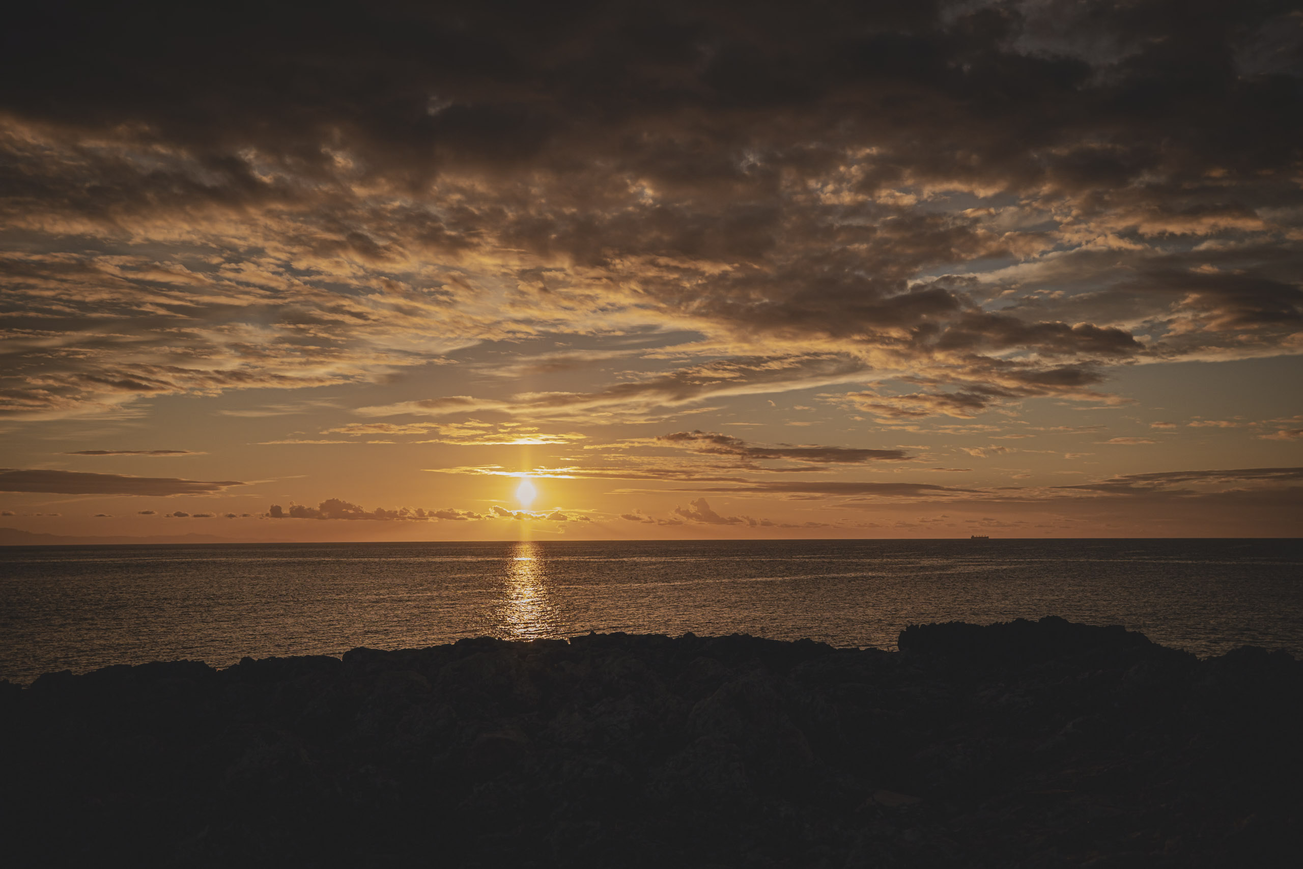Ein dramatischer Sonnenuntergang über dem Meer, die Sonne steht tief am Horizont und wirft einen warmen Schein unter einen Himmel voller dunkler, strukturierter Wolken, betrachtet von einer felsigen Küste. © Fotografie Tomas Rodriguez