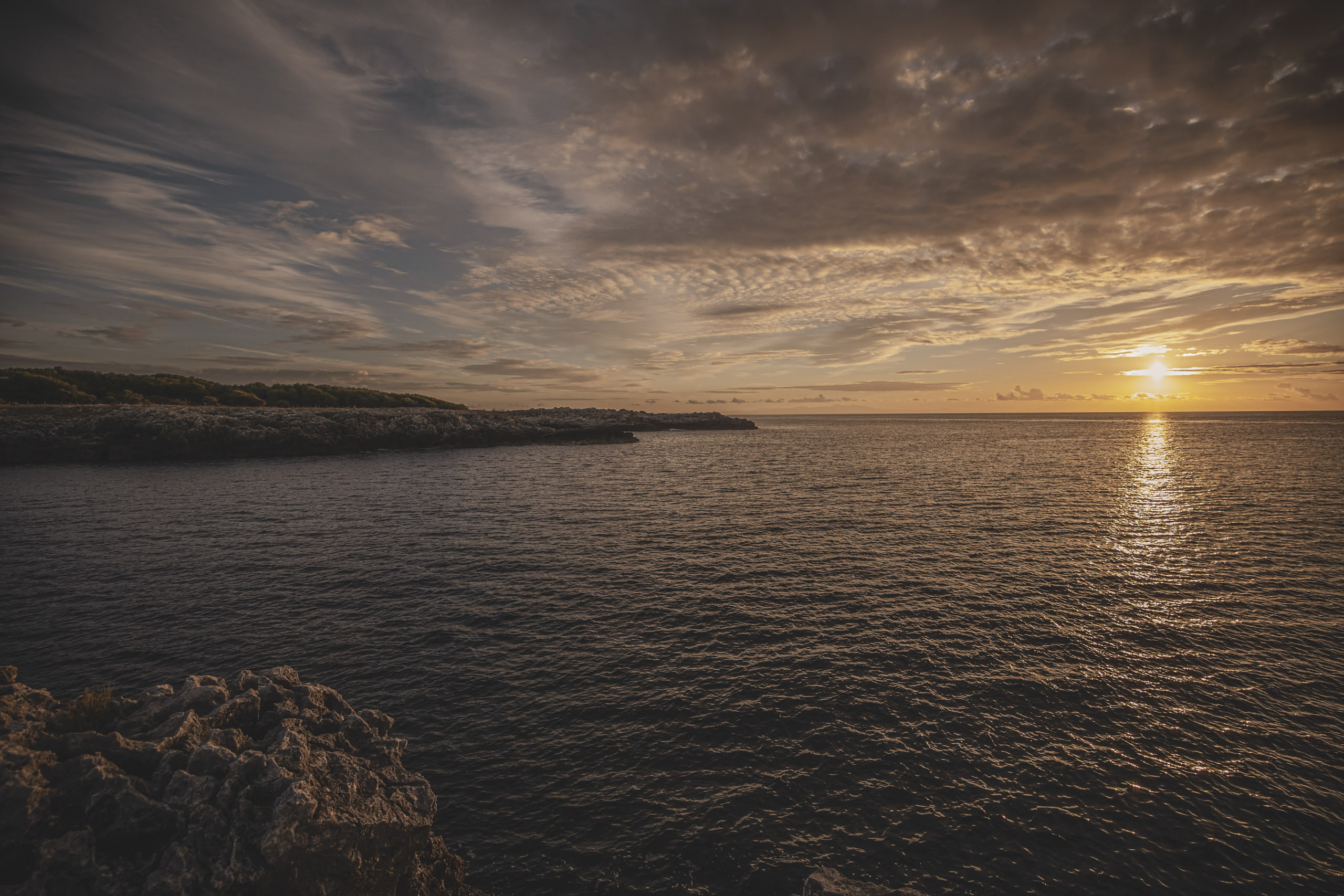 Sonnenuntergang über einem ruhigen Meer, wobei die Sonne einen goldenen Pfad auf das Wasser wirft. Dunkle, strukturierte Wolken schweben über einer zerklüfteten Küste. © Fotografie Tomas Rodriguez