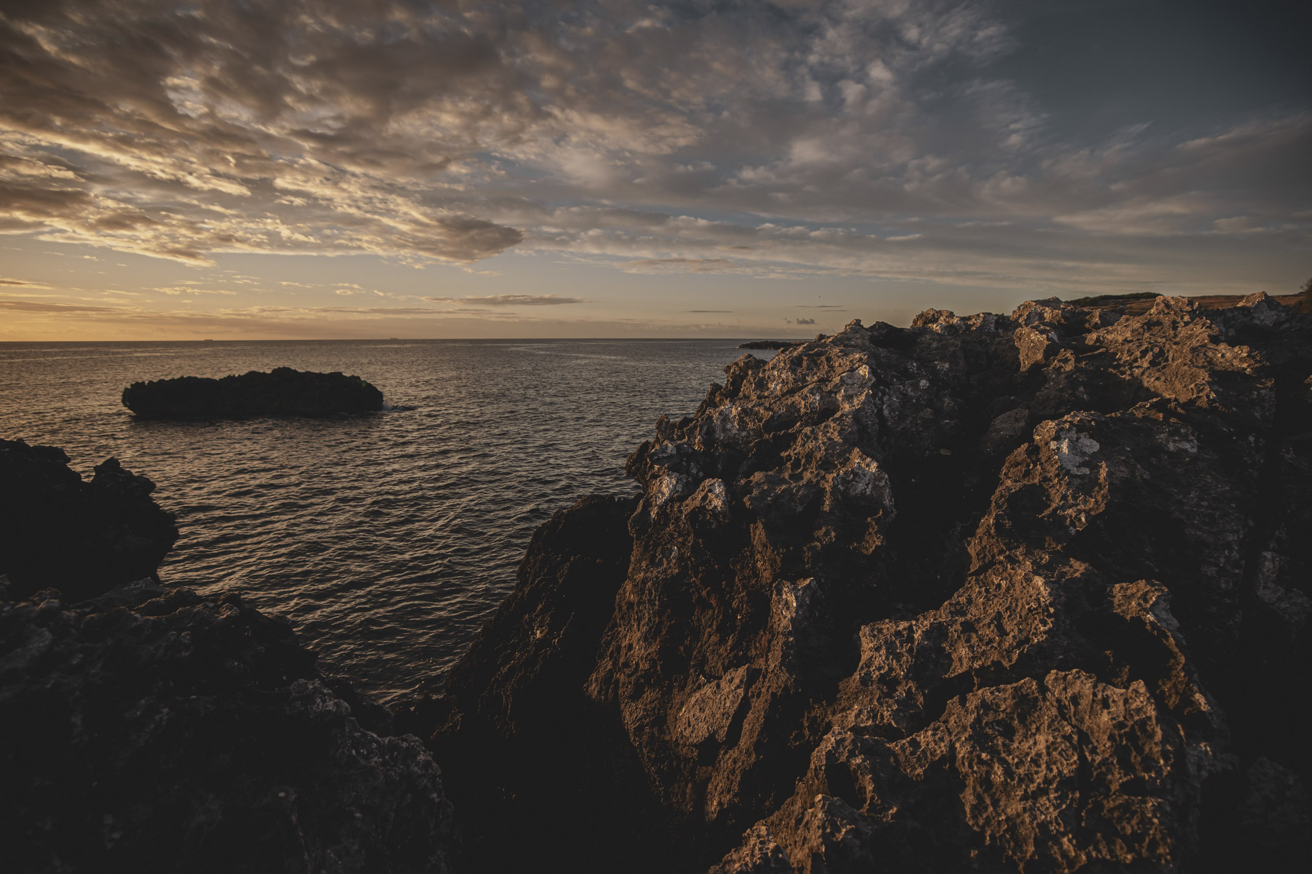 Eine dramatische Küstenszene bei Sonnenuntergang mit schroffen Klippen und ruhigem Meer unter einem bewölkten Himmel, durch den goldenes Sonnenlicht durchdringt. © Fotografie Tomas Rodriguez