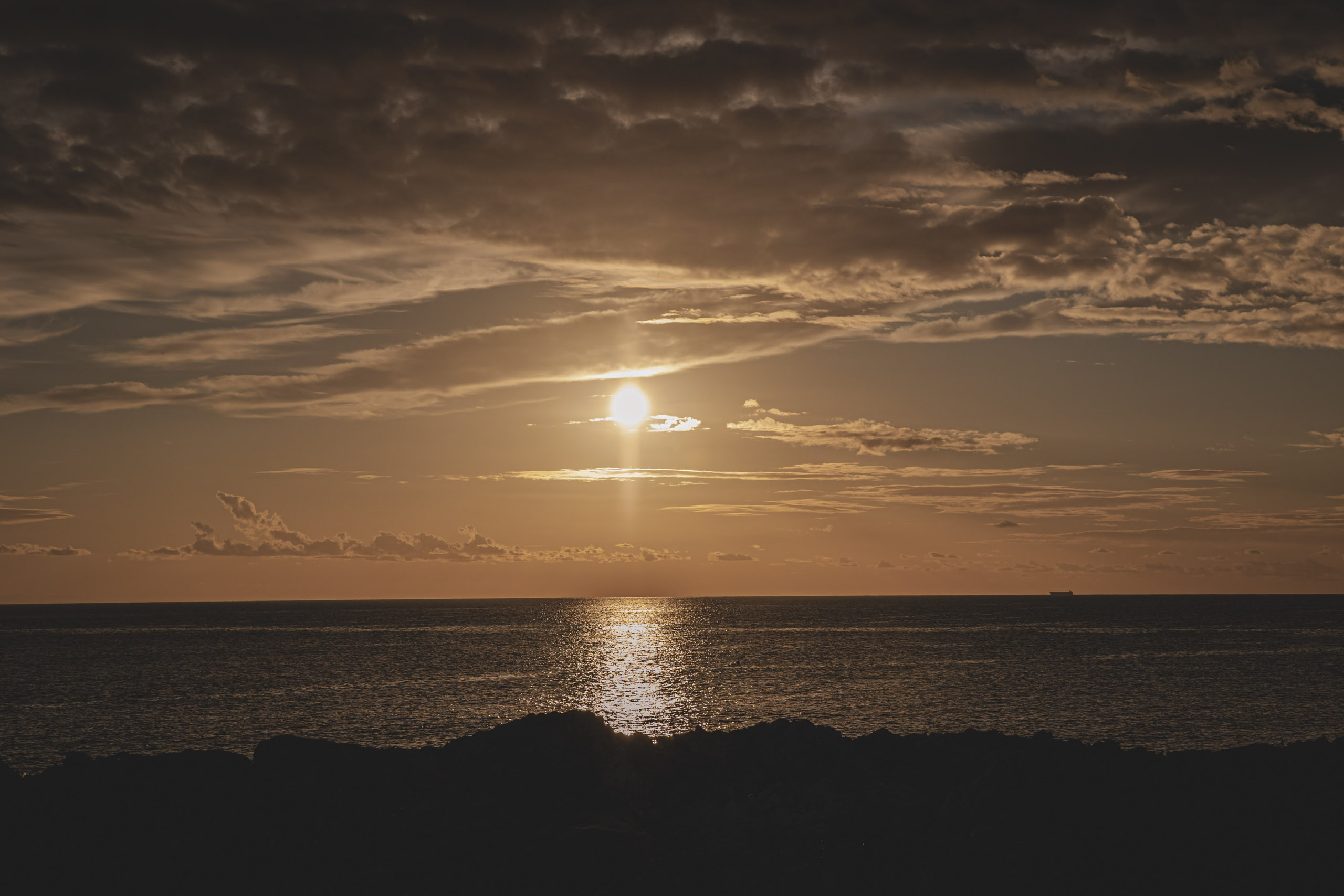 Die Sonne geht über dem Ozean unter und wirft einen goldenen Widerschein auf das Wasser, mit einer felsigen Küstenlinie im Vordergrund und einem entfernten Schiff am Horizont unter einem teilweise bewölkten Himmel. © Fotografie Tomas Rodriguez