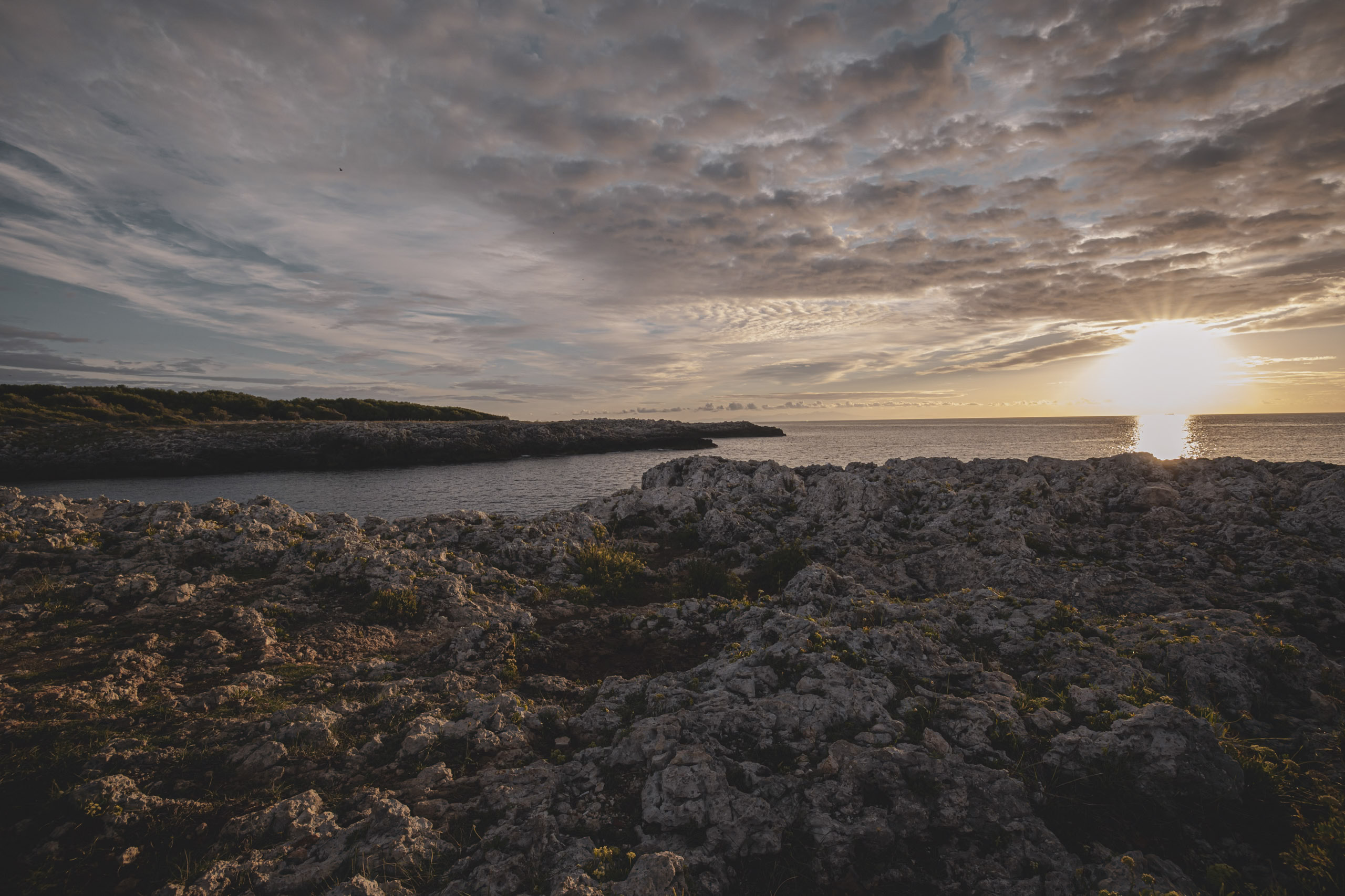 Ein malerischer Sonnenuntergang über einer zerklüfteten Küste, mit einem dramatischen Himmel mit vereinzelten Wolken und der Sonne nahe dem Horizont, die einen warmen Schein über die felsige Landschaft wirft. © Fotografie Tomas Rodriguez