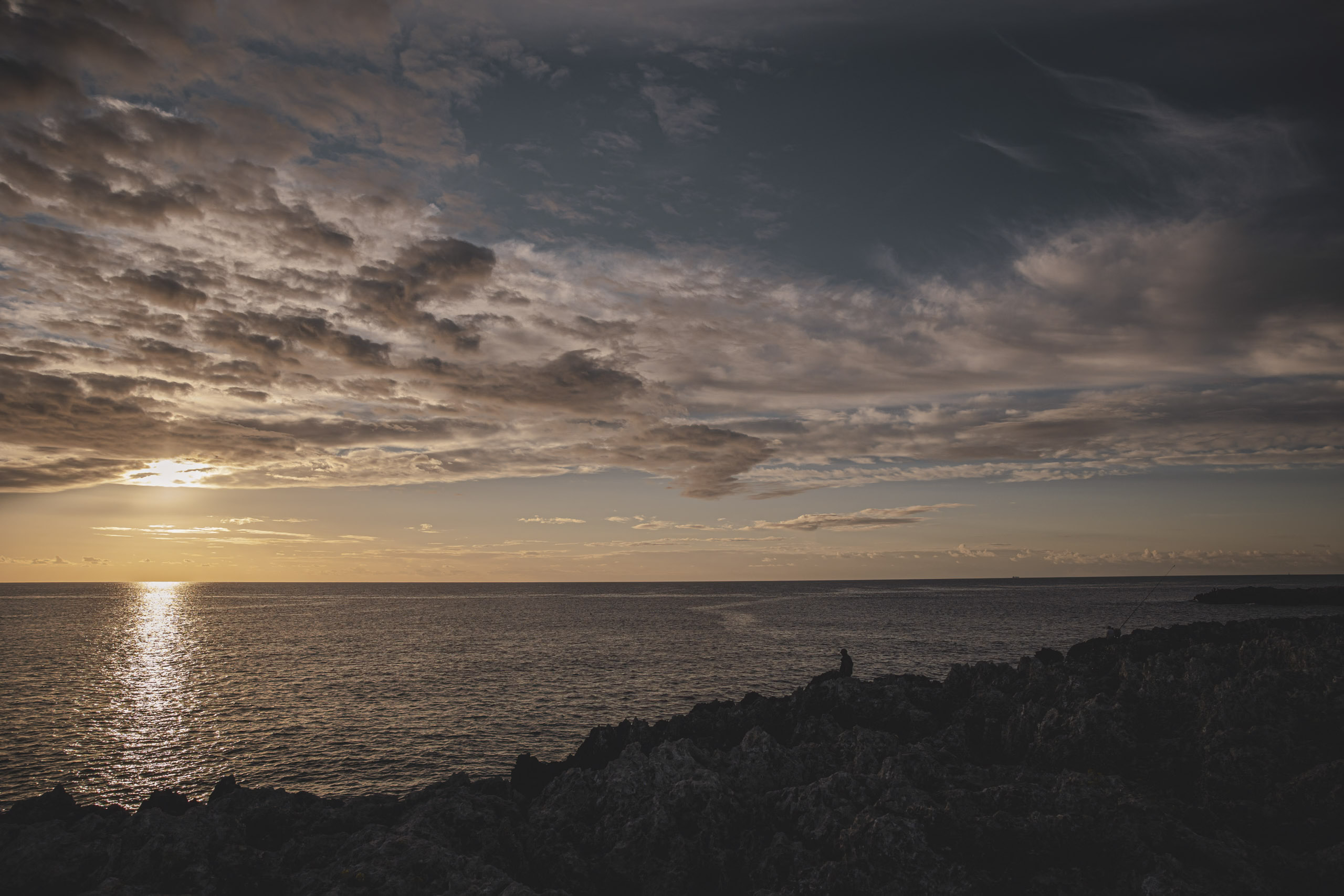 Ein ruhiger Sonnenuntergang über dem Meer, von einer felsigen Küste aus betrachtet. Der Himmel ist mit Wolken übersät, die das warme Sonnenlicht reflektieren. Am Wasserrand steht eine einsame Gestalt als Silhouette. © Fotografie Tomas Rodriguez