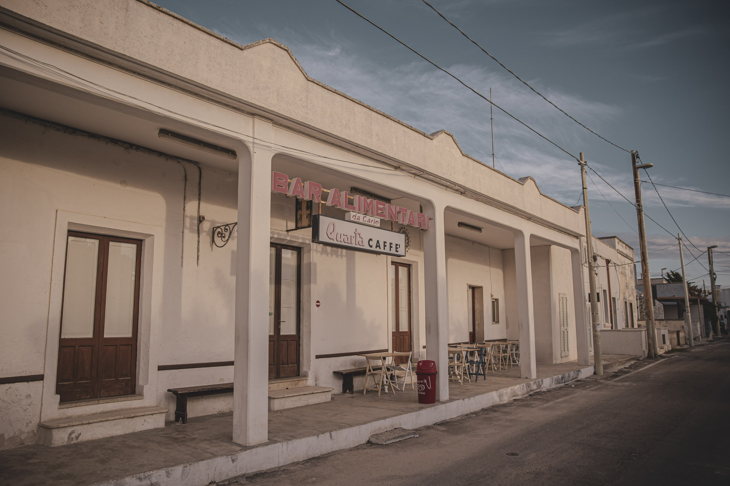 Eine ruhige Straßenszene mit einem einstöckigen Gebäude und einem Schild mit der Aufschrift „Bar Alimentari“, das auf eine lokale Café-Bar in rustikaler Umgebung unter einem klaren Himmel in der Abenddämmerung hinweist. © Fotografie Tomas Rodriguez