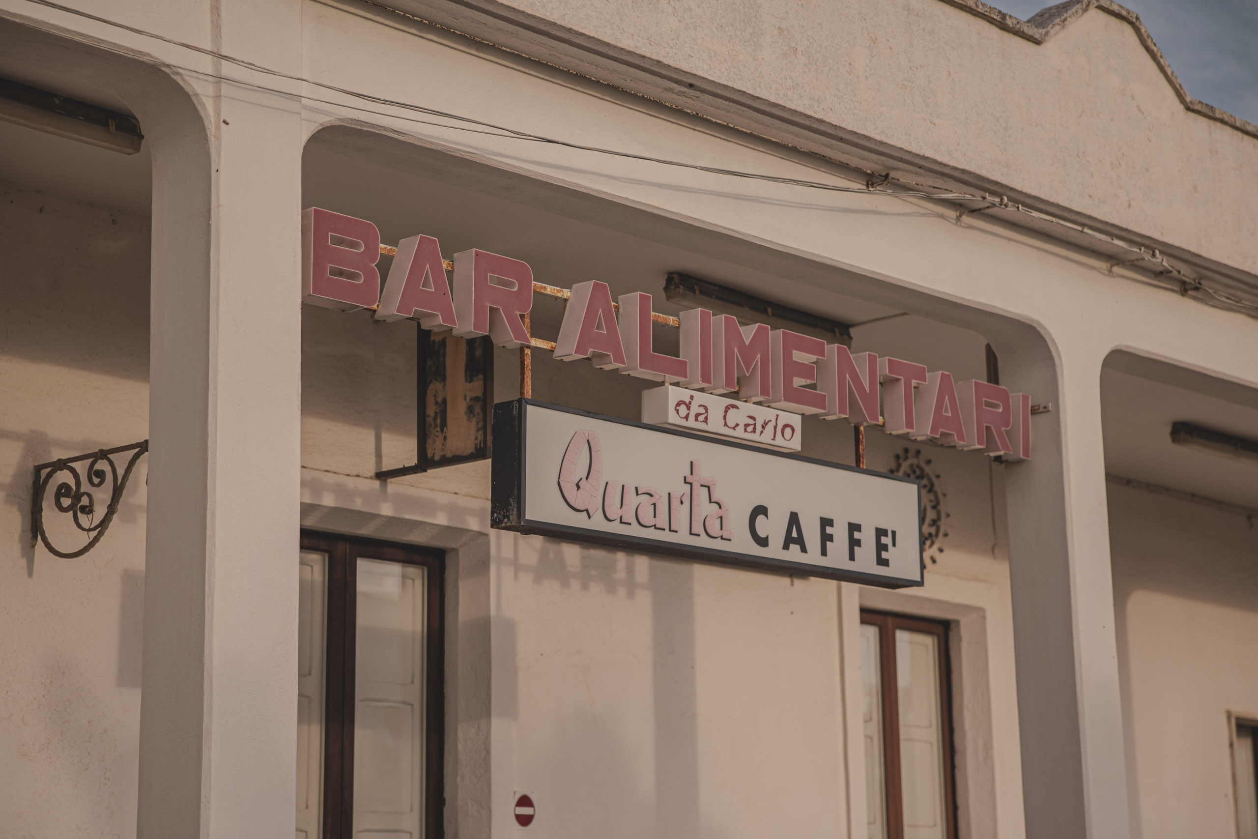 Schilder mit der Aufschrift „Bar Alimentari da Carlo“ und „Qualità Caffè“ an einem beigefarbenen Gebäude mit dekorativen Schmiedearbeiten unter klarem Himmel. © Fotografie Tomas Rodriguez