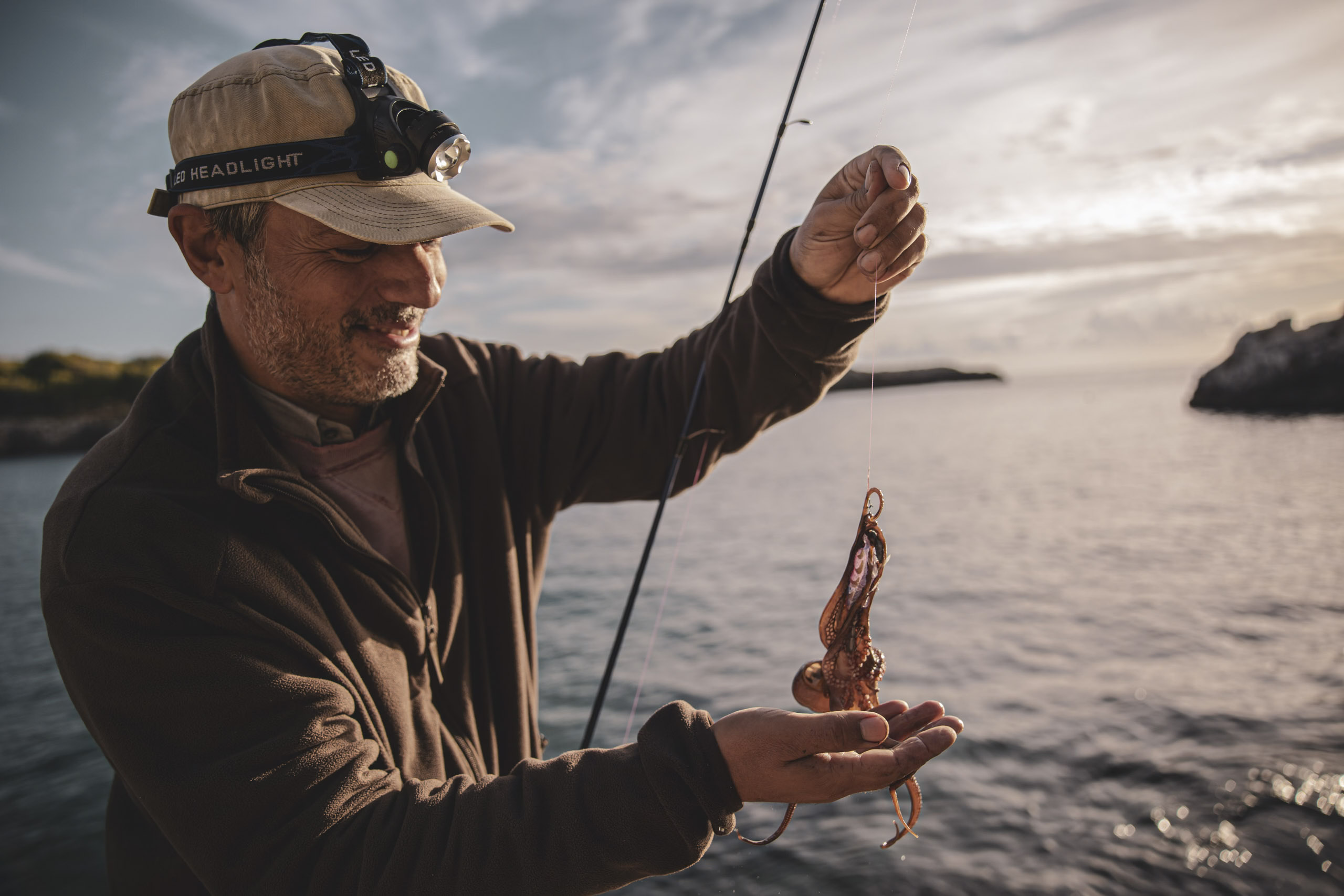 Ein Mann mit Stirnlampe und Mütze lächelt, während er eine Angelrute hält und vor der Kulisse eines Sonnenuntergangs an der Küste einen Tintenfisch am Haken untersucht. © Fotografie Tomas Rodriguez