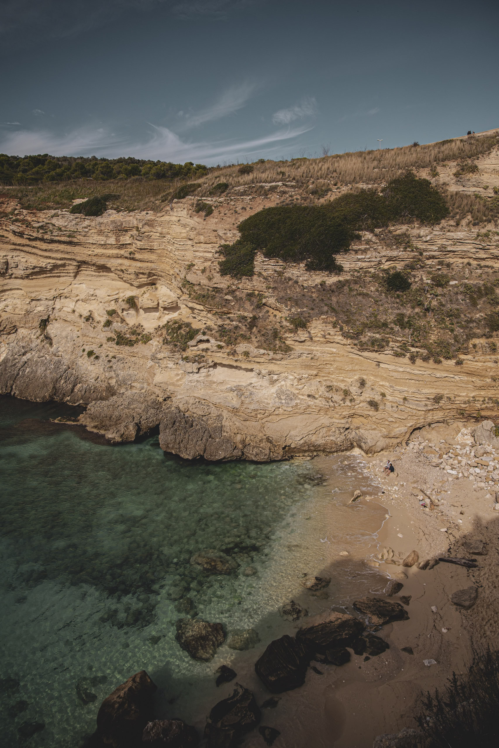 Ein malerischer Blick auf eine felsige Bucht mit klarem, türkisfarbenem Wasser, umgeben von geschichteten Klippen und spärlicher Vegetation unter einem klaren, blauen Himmel. © Fotografie Tomas Rodriguez