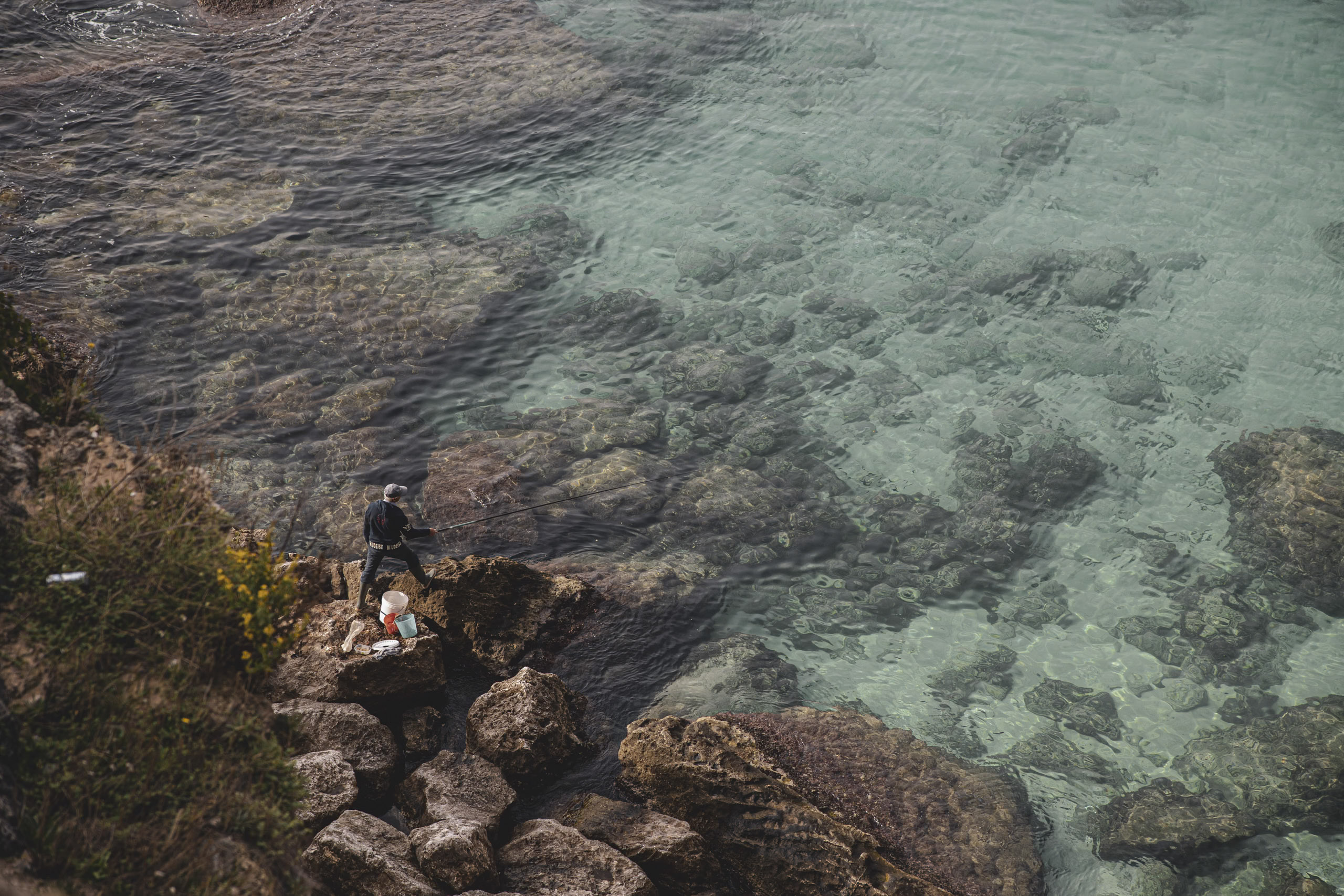 Zwei Personen angeln vor einer felsigen Küste. Eine Person sitzt, die andere steht. Das klare, türkisfarbene Wasser gibt den Blick auf untergetauchte Felsen und sanfte Wellen frei. © Fotografie Tomas Rodriguez