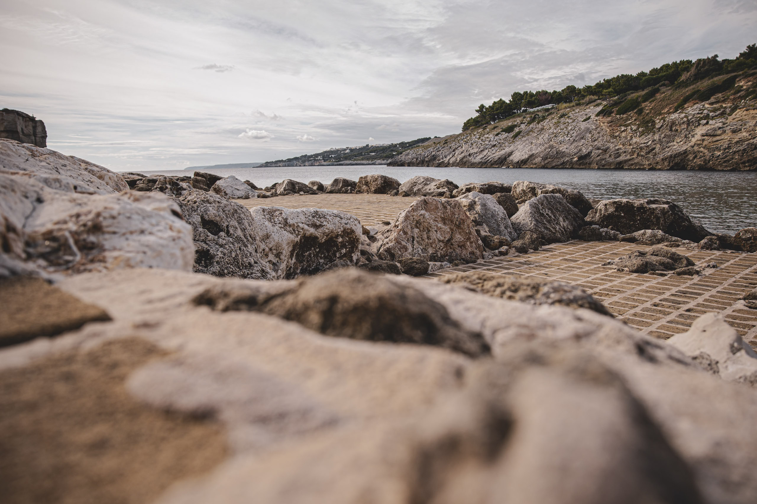 Raue, strukturierte Felsen im Vordergrund mit einem Steinpfad, der unter einem teilweise bewölkten Himmel zu einem ruhigen Meer führt. In der Ferne sind Hügel sichtbar, die eine ruhige Küstenlandschaft vervollständigen. © Fotografie Tomas Rodriguez