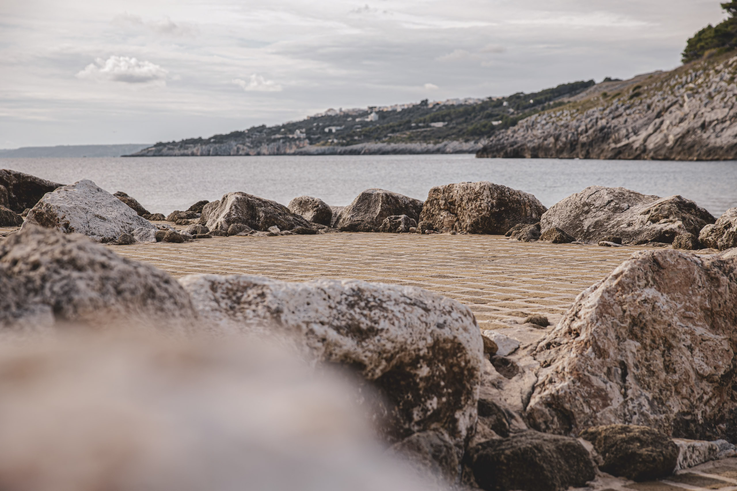 Ein von Felsen eingerahmter Küstenblick. Im Vordergrund ist ein gefliester Weg zu sehen, der zum Meer führt. Im Hintergrund sind sanfte Hügel und ein klarer Himmel zu sehen. © Fotografie Tomas Rodriguez