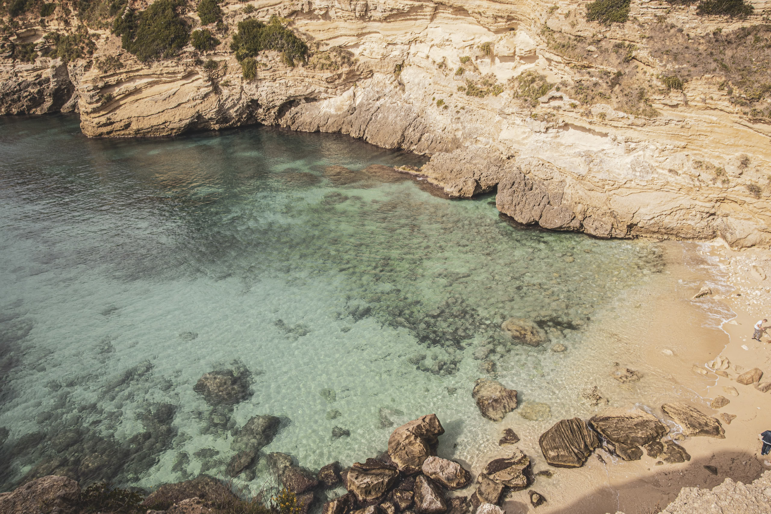 Luftaufnahme einer abgeschiedenen Bucht mit türkisfarbenem Wasser, begrenzt von Felsklippen, mit einem kleinen Sandstrand und verstreuten Felsbrocken entlang der Uferlinie. © Fotografie Tomas Rodriguez