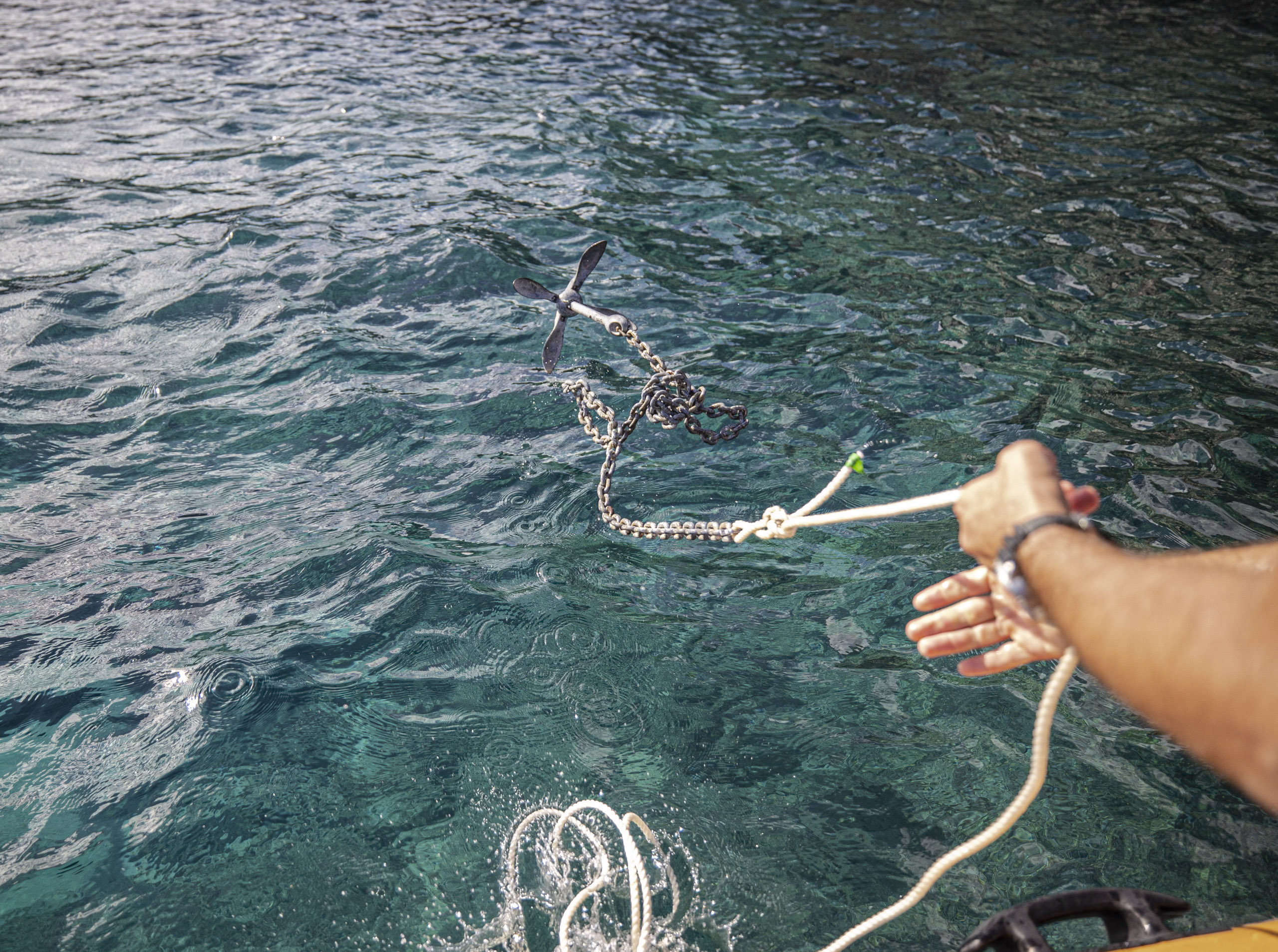 Die Hand einer Person hält ein Seil, das an einem Metallanker befestigt ist, der teilweise in klares, türkisfarbenes Wasser eingetaucht ist, wobei sich das Sonnenlicht auf der Meeresoberfläche spiegelt. © Fotografie Tomas Rodriguez