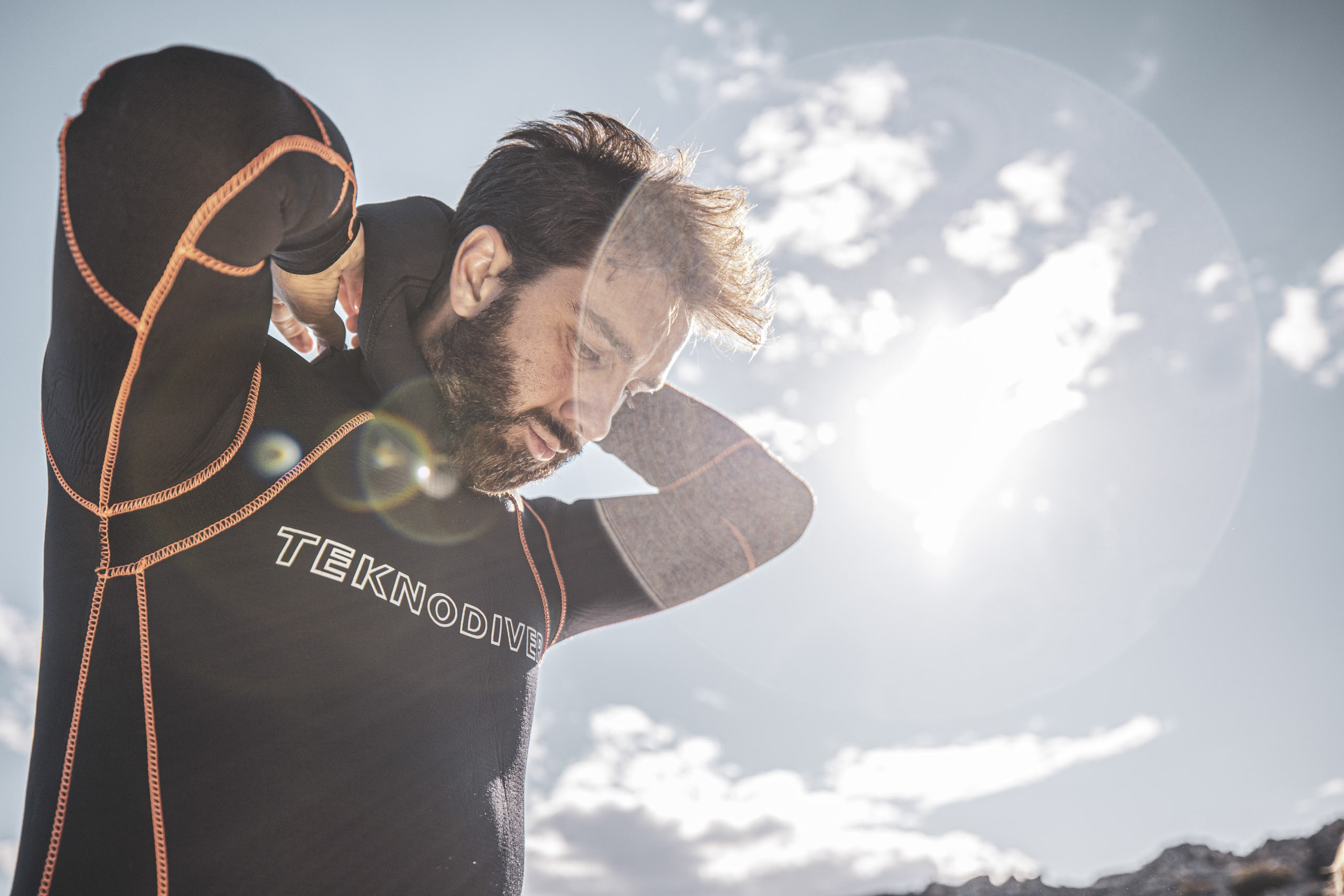 Ein Mann richtet seinen Neoprenanzug in der hellen Sonne, mit leichtem Linsenreflexion und bewölktem Himmel im Hintergrund, was einen aktiven Moment im Freien hervorhebt. © Fotografie Tomas Rodriguez