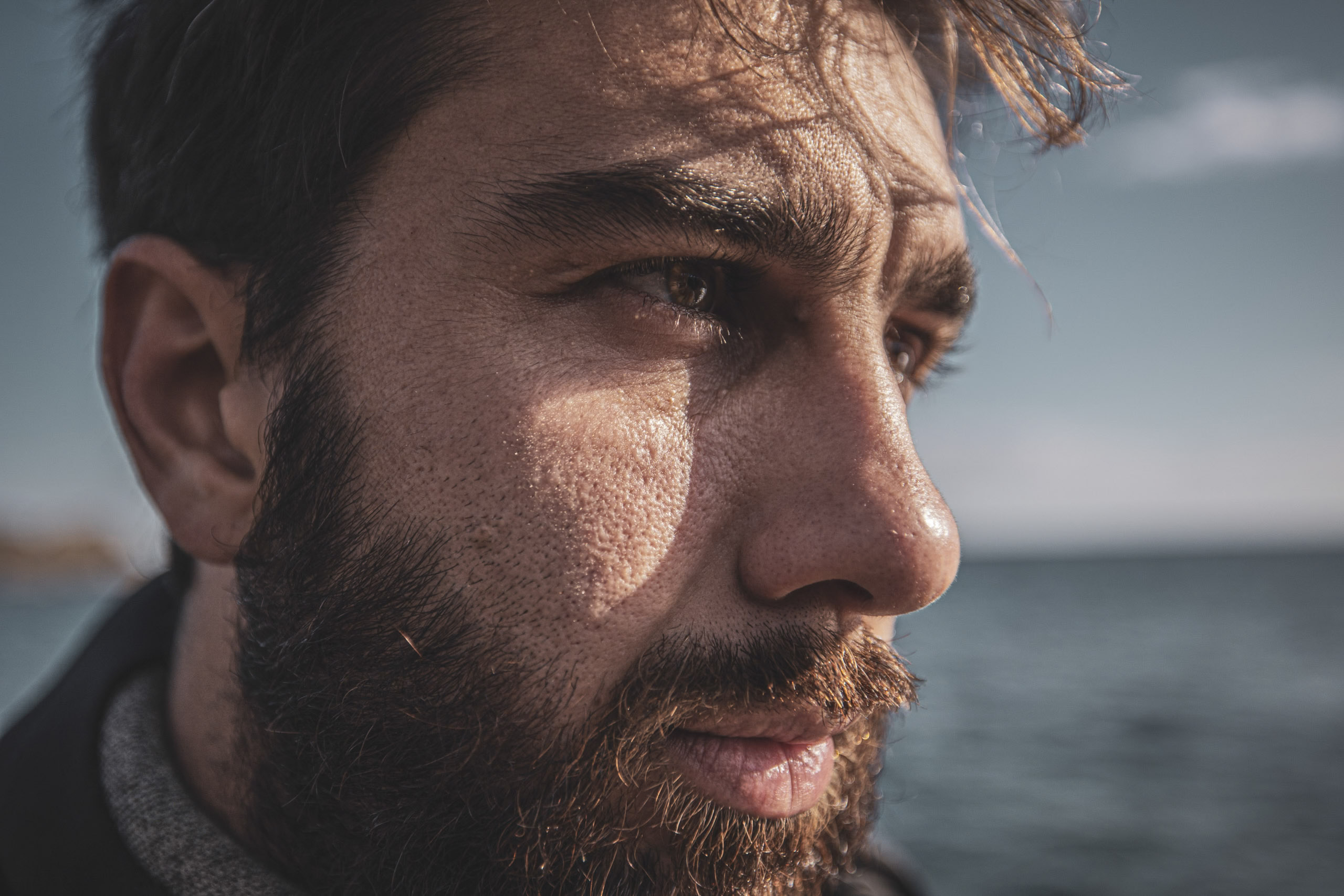 Nahaufnahme eines Mannes mit Bart, der in die Ferne blickt. Profilaufnahme im Freien mit dem Meer im Hintergrund, die detaillierte Gesichtszüge und den intensiven Blick hervorhebt. © Fotografie Tomas Rodriguez
