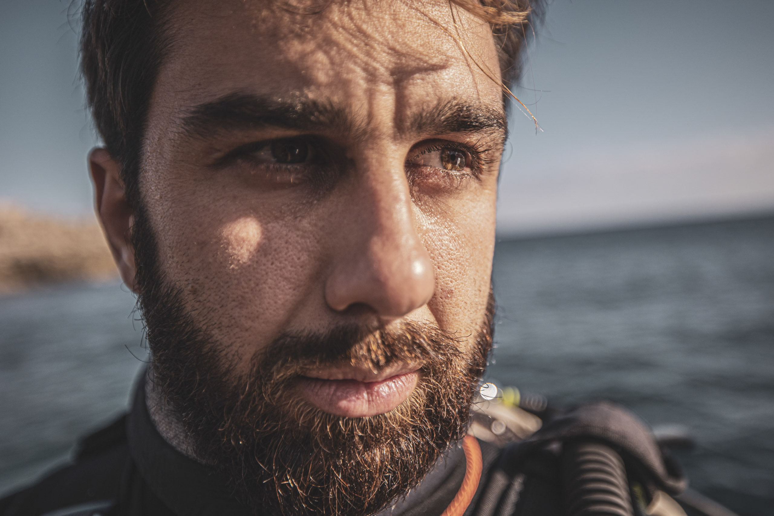 Nahaufnahme eines bärtigen Mannes mit nachdenklichem Gesichtsausdruck und Taucherausrüstung. Im Hintergrund sind das Meer und eine felsige Küstenlinie leicht unscharf. © Fotografie Tomas Rodriguez