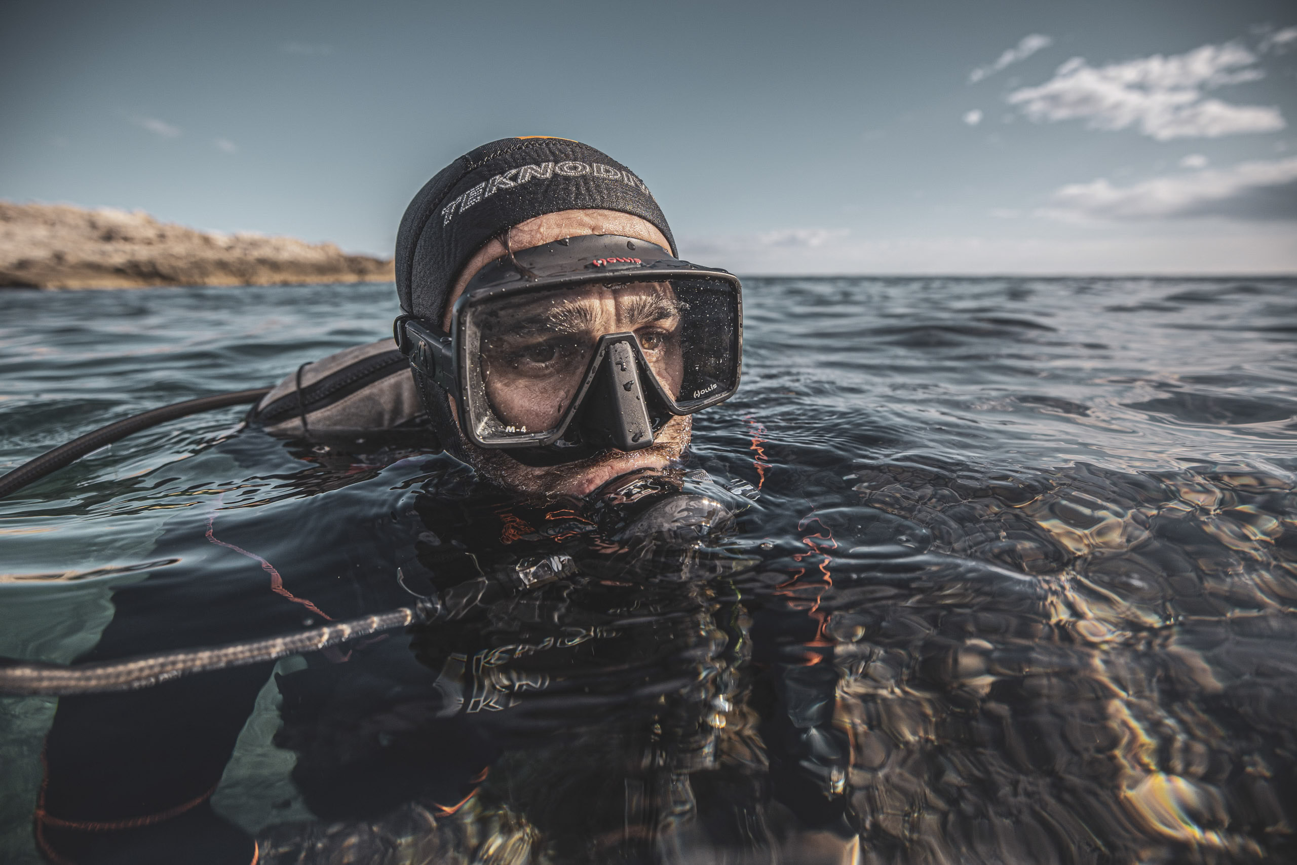 Ein teilweise im Meerwasser eingetauchter Taucher mit Maske und Schnorchel blickt mit ruhigem Gesichtsausdruck aufmerksam in die Kamera, im Hintergrund sind felsige Küsten zu sehen. © Fotografie Tomas Rodriguez