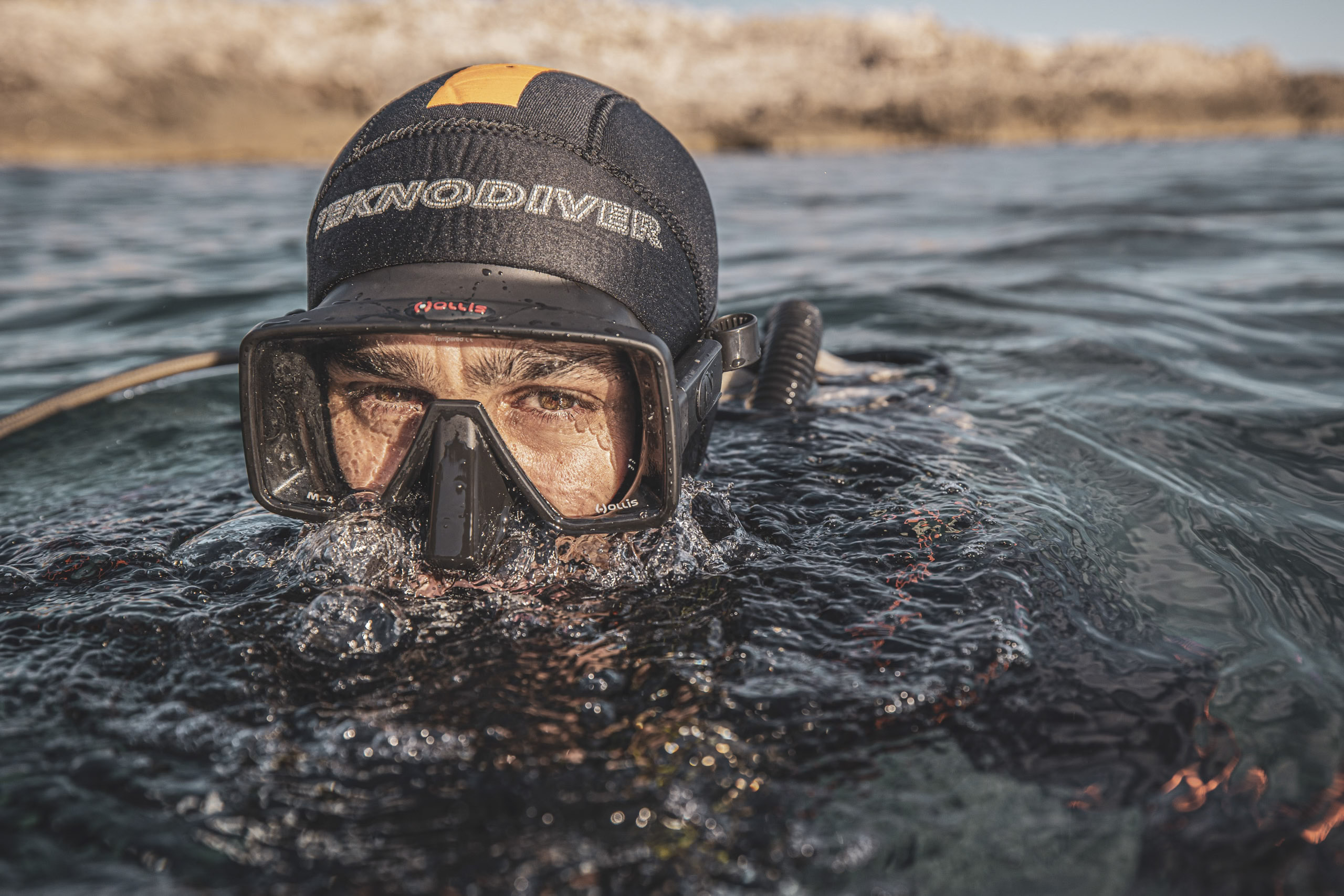 Nahaufnahme einer Person mit Tauchermaske und Schnorchel, die teilweise unter Wasser ist. Auf ihrer Neoprenhaube steht „Skinodiver“. Im Hintergrund ist eine verschwommene Wasserlandschaft zu sehen. © Fotografie Tomas Rodriguez