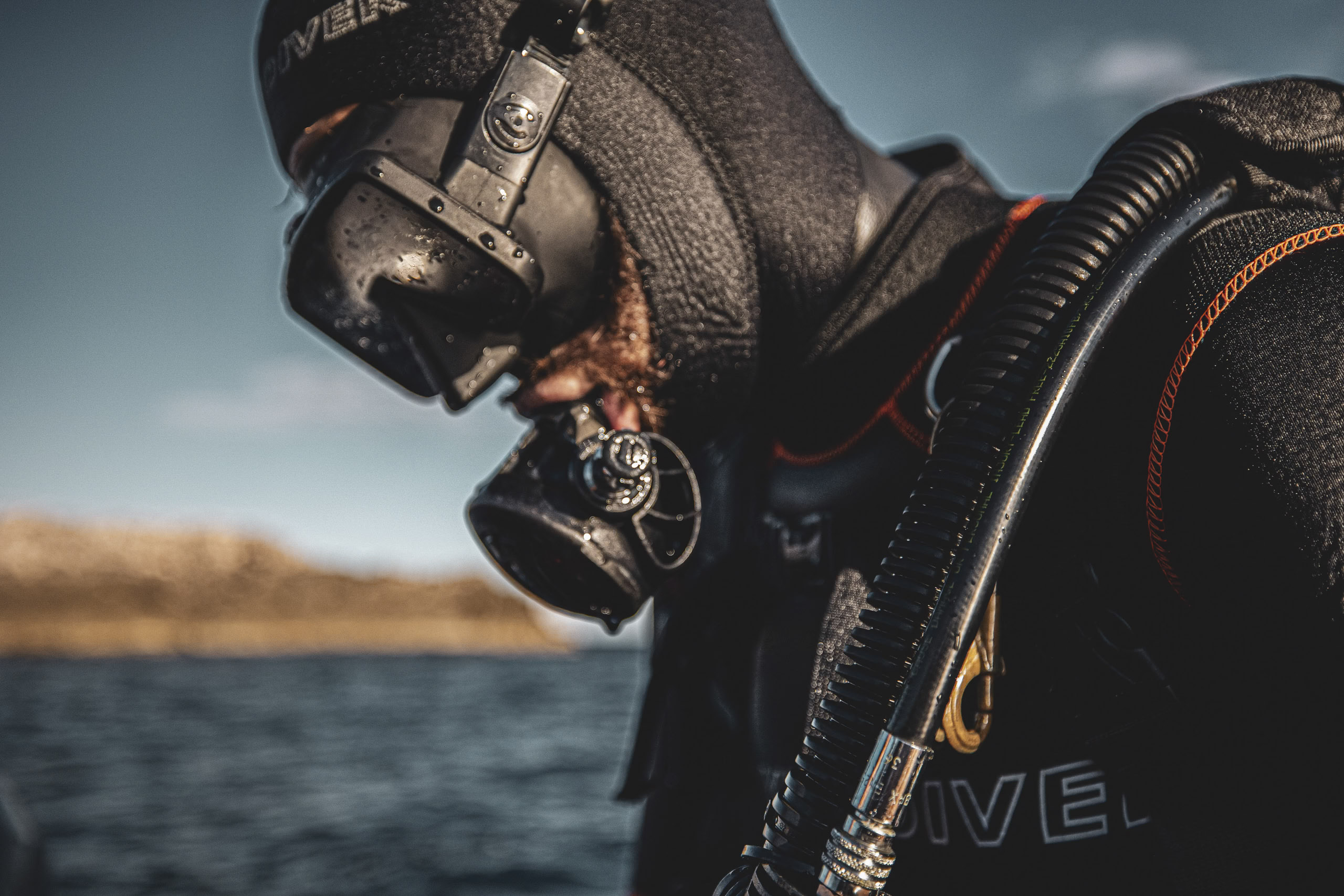 Nahaufnahme eines Tauchers, der eine Vollgesichtsmaske und Tauchausrüstung einschließlich Trockenanzug trägt, mit klarem blauen Wasser und Sonnenlicht im Hintergrund. © Fotografie Tomas Rodriguez