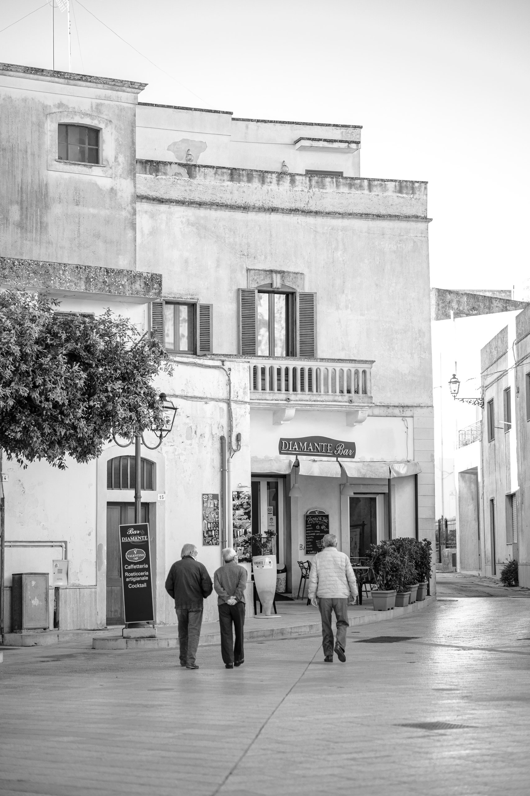 Drei ältere Männer gehen gemeinsam durch eine malerische Straße, die von traditionellen Gebäuden gesäumt ist, darunter ein Geschäft namens „Diamante Shoes“. Die Szene fängt eine heitere Altstadtatmosphäre ein. © Fotografie Tomas Rodriguez