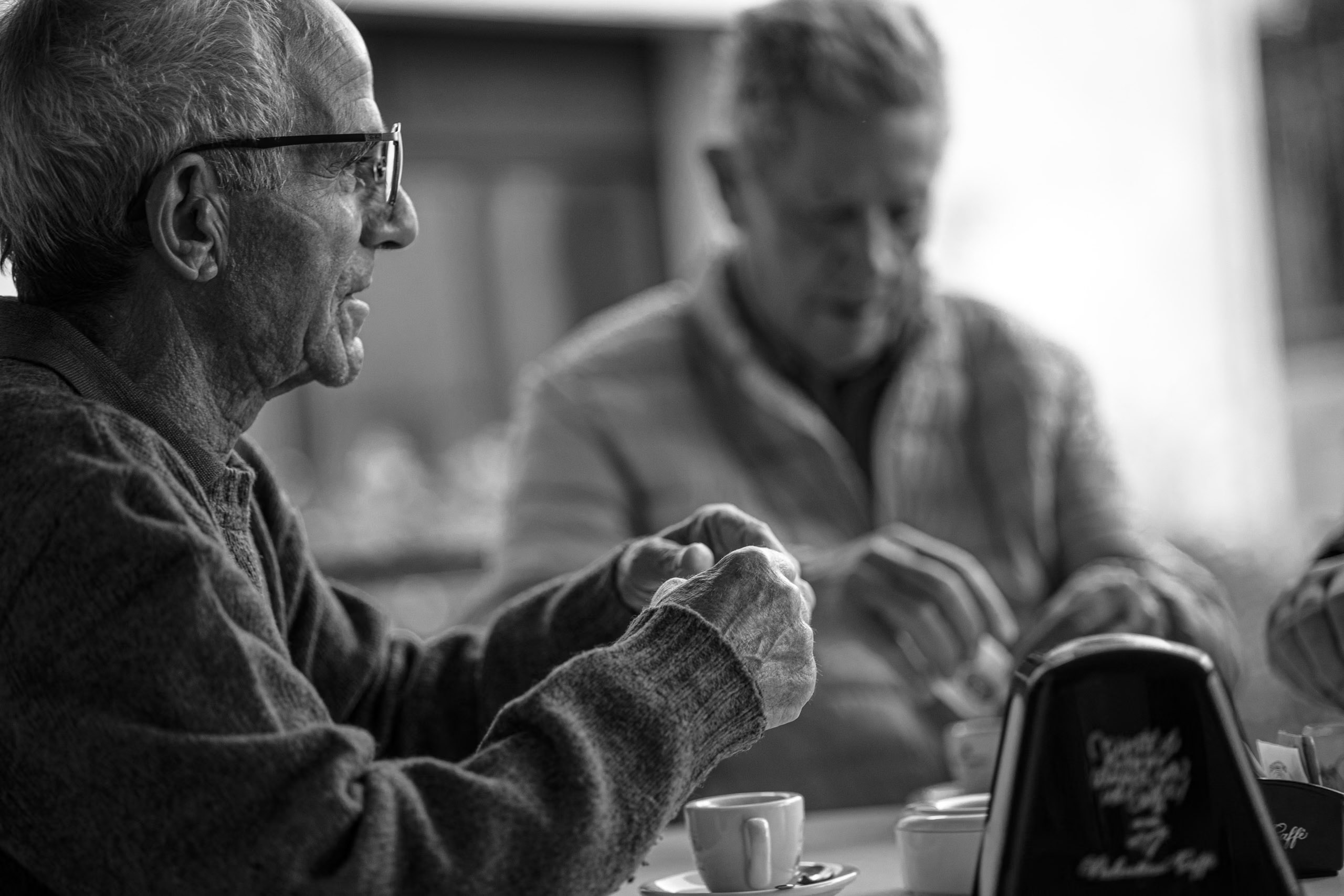 Zwei ältere Männer sind an einem Cafétisch in ein angeregtes Gespräch verwickelt. Einer gestikuliert mit den Händen, der andere ist auf das Mischen der Karten konzentriert. Auf dem Tisch liegen eine kleine Kaffeetasse und eine Zigarettenpackung. © Fotografie Tomas Rodriguez
