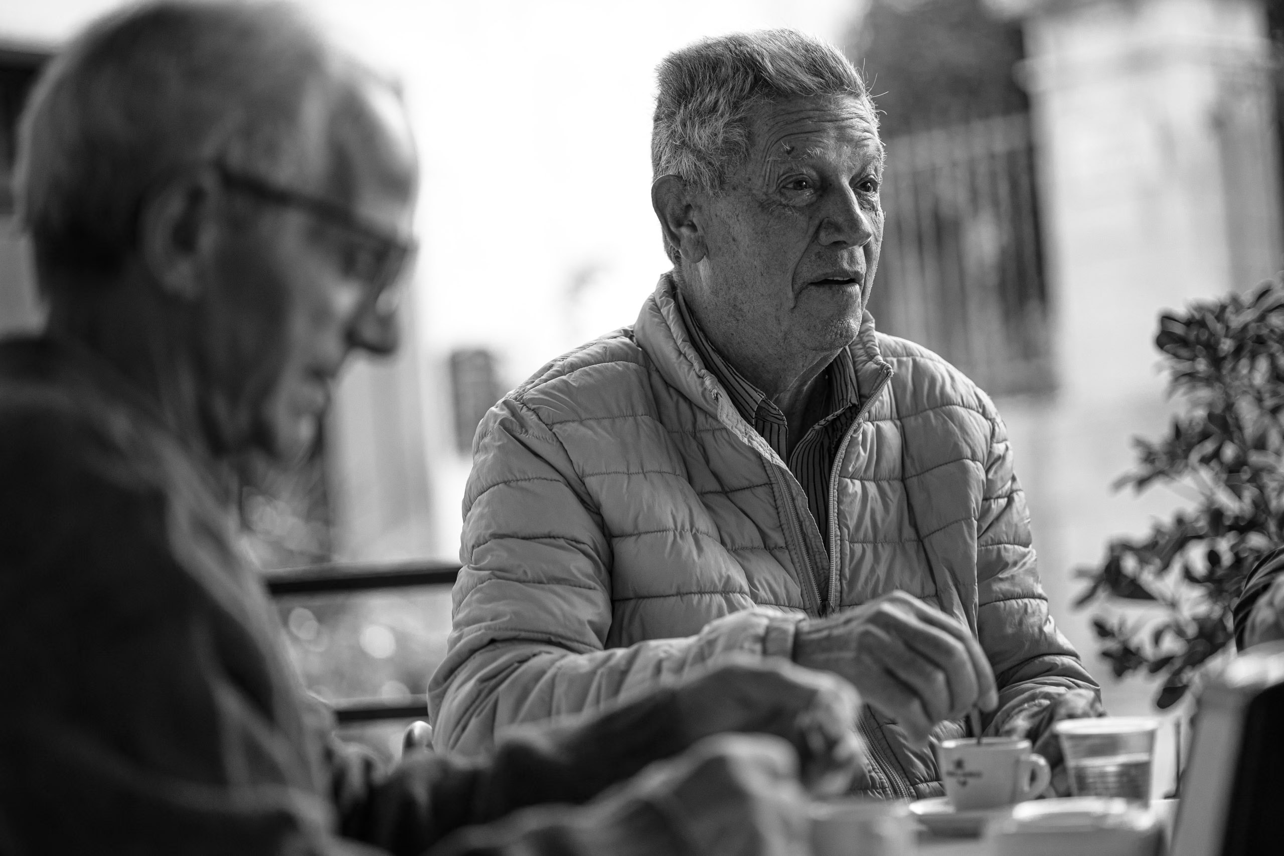 Zwei ältere Männer sitzen an einem Tisch und unterhalten sich. Ein Mann konzentriert sich auf seinen Begleiter, der zu sprechen scheint, während die entspannte Atmosphäre im Freien herrscht. © Fotografie Tomas Rodriguez
