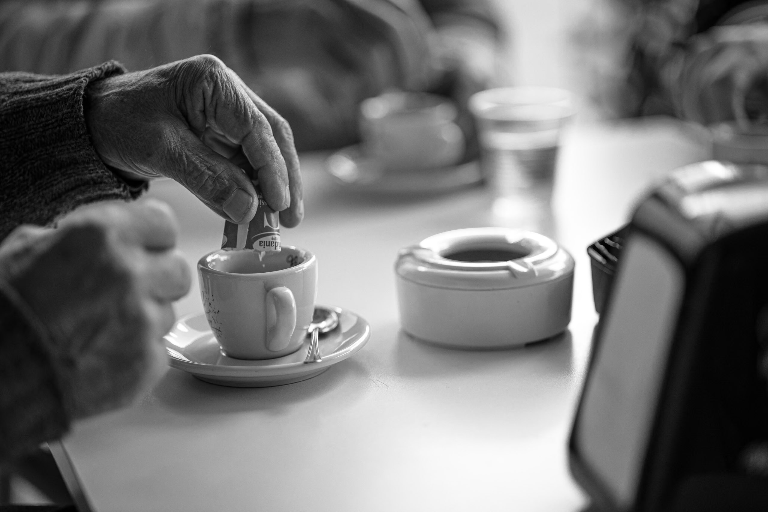 Die Hände einer älteren Person rühren mit einem Löffel eine kleine weiße Tasse Espresso um, die auf einer Untertasse neben einem Aschenbecher auf einem Tisch steht, aufgenommen in Schwarzweiß. © Fotografie Tomas Rodriguez