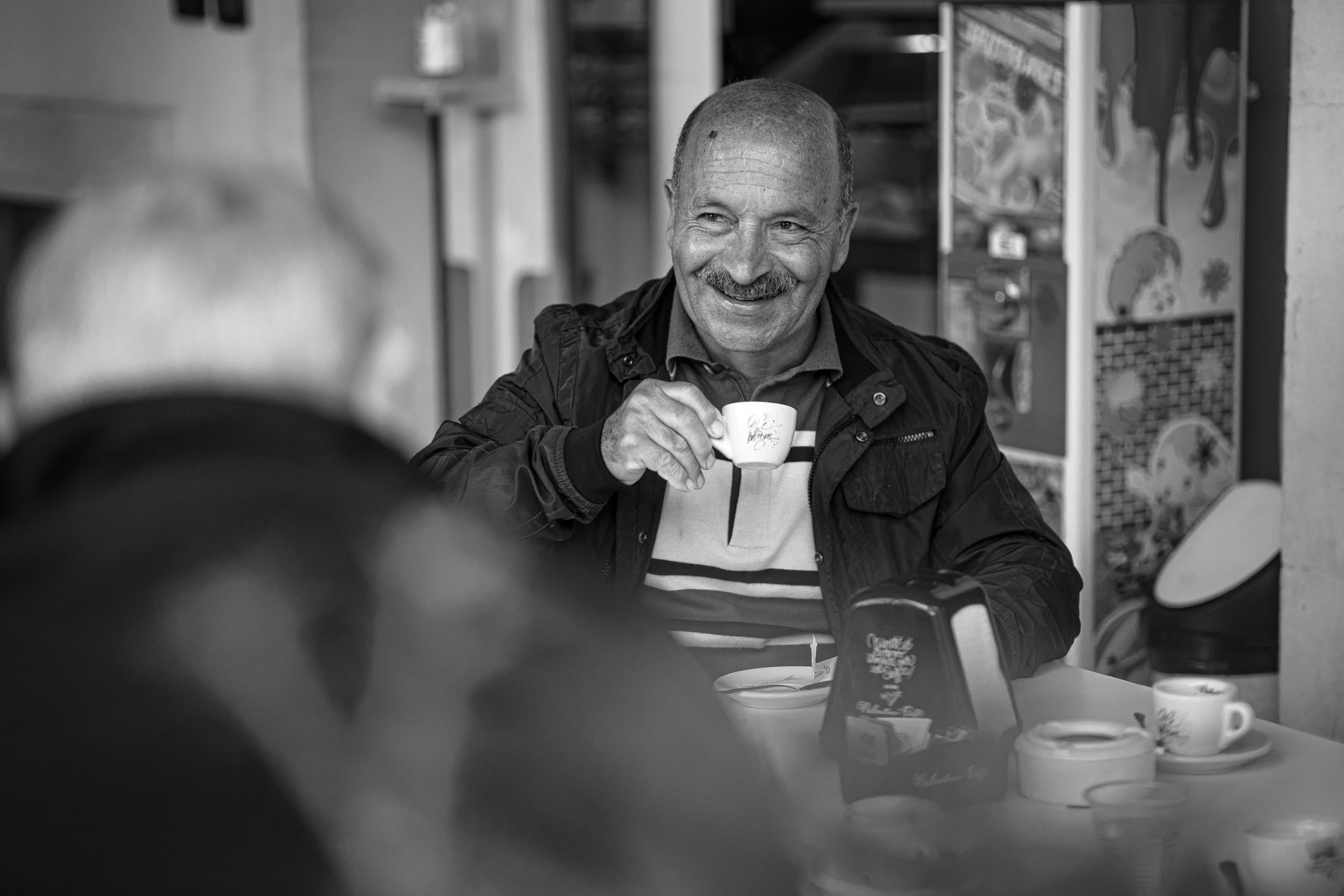 Ein fröhlicher älterer Mann hält in einem Café eine Kaffeetasse und lächelt einer anderen Person am anderen Ende des Tisches herzlich zu. Das Bild ist schwarz-weiß und hebt das Lachen und die entspannte Atmosphäre hervor. © Fotografie Tomas Rodriguez