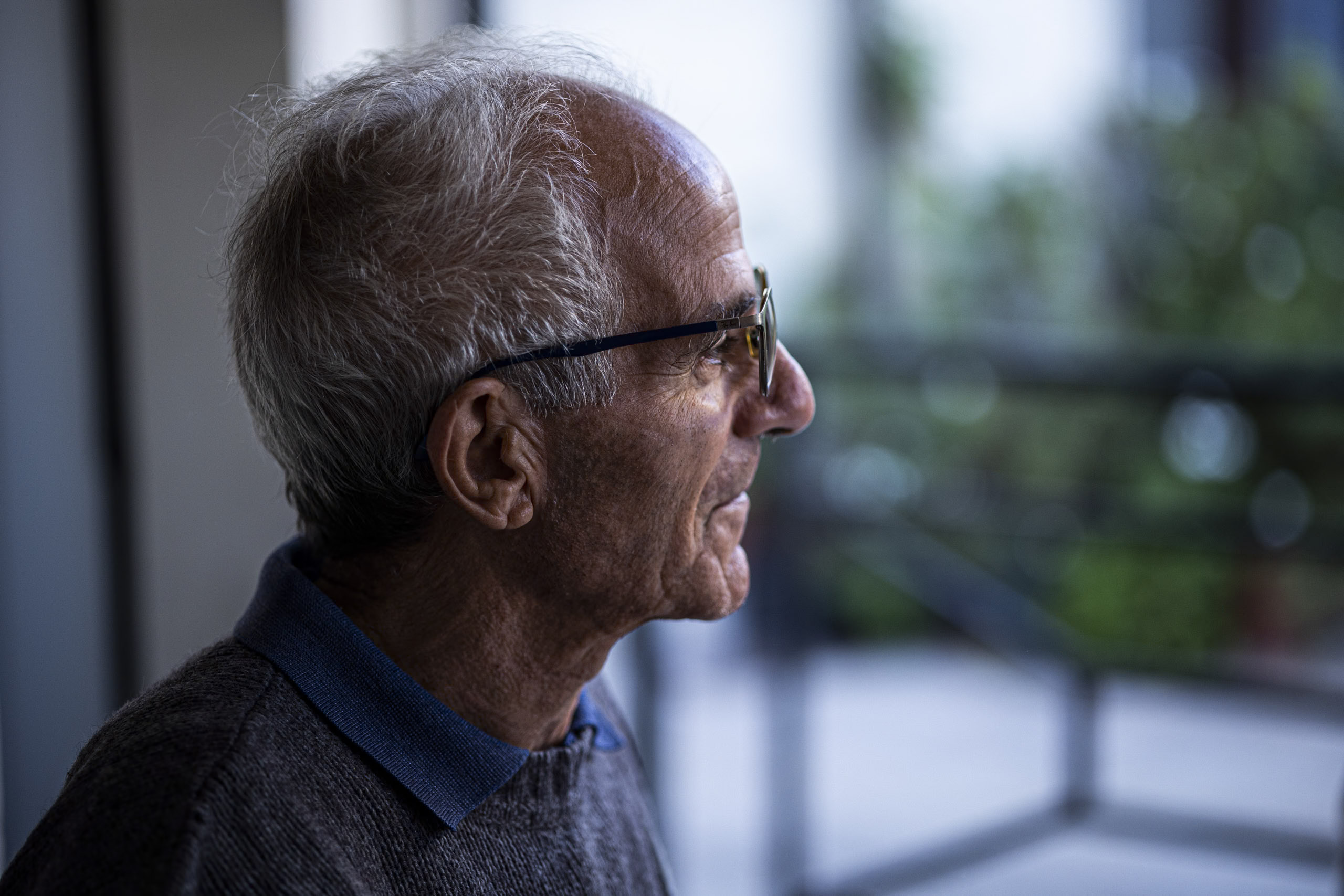 Profilansicht eines älteren Mannes mit weißem Haar und Brille, der mit nachdenklichem Gesichtsausdruck aus dem Fenster blickt. Weicher Fokus auf grünem, unscharfem Hintergrund. © Fotografie Tomas Rodriguez
