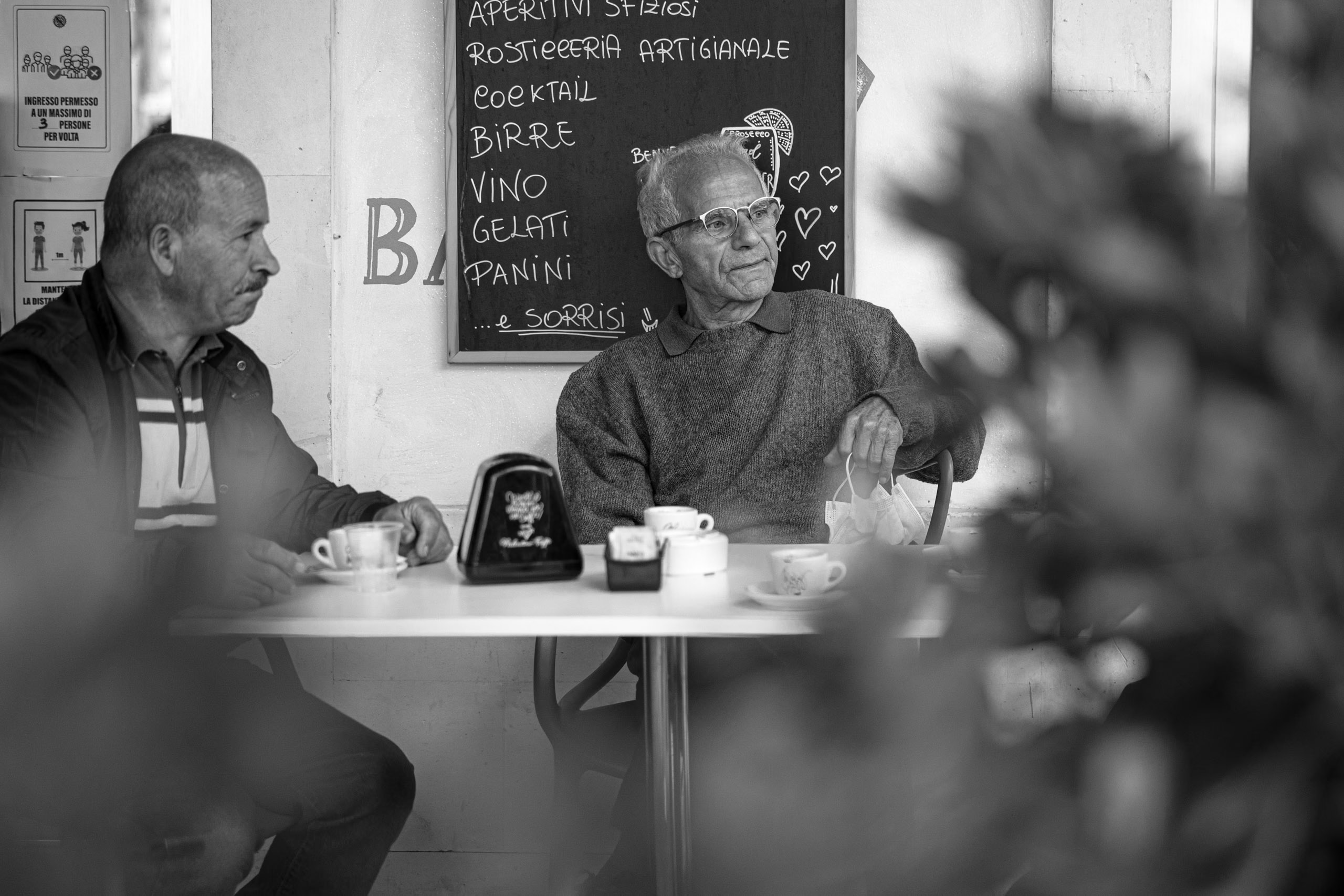 Zwei Männer sitzen an einem kleinen Cafétisch, einer gestikuliert mitten im Gespräch. Im Vordergrund ist eine verschwommene Pflanze zu sehen, im Hintergrund ist eine Menütafel zu erkennen. Schwarzweißfoto. © Fotografie Tomas Rodriguez