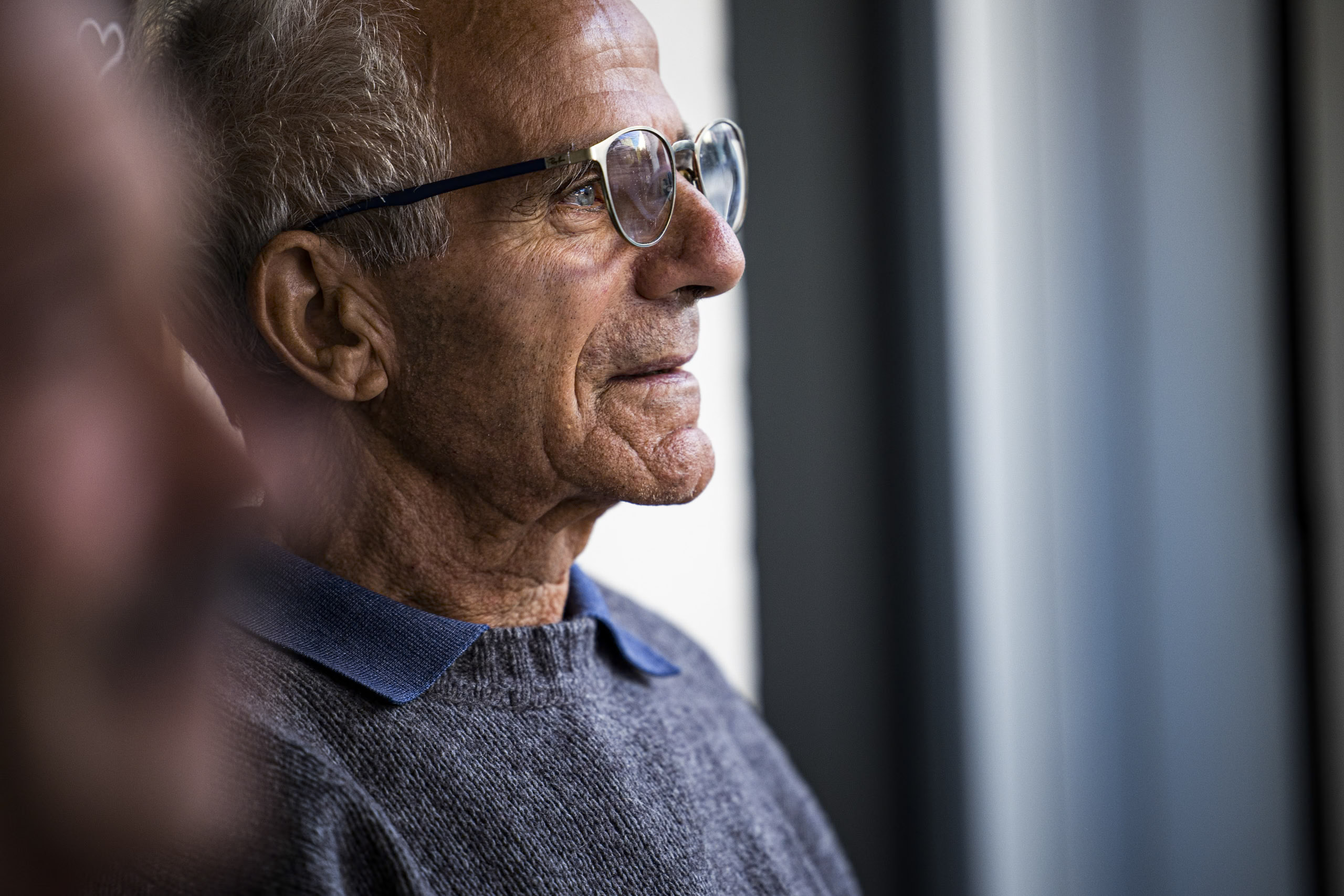 Profil eines älteren Mannes mit Brille, der in Gedanken versunken aus dem Fenster blickt. Er trägt einen Pullover und hat einen nachdenklichen Gesichtsausdruck, der ein Gefühl der Kontemplation oder Erinnerung vermittelt. © Fotografie Tomas Rodriguez