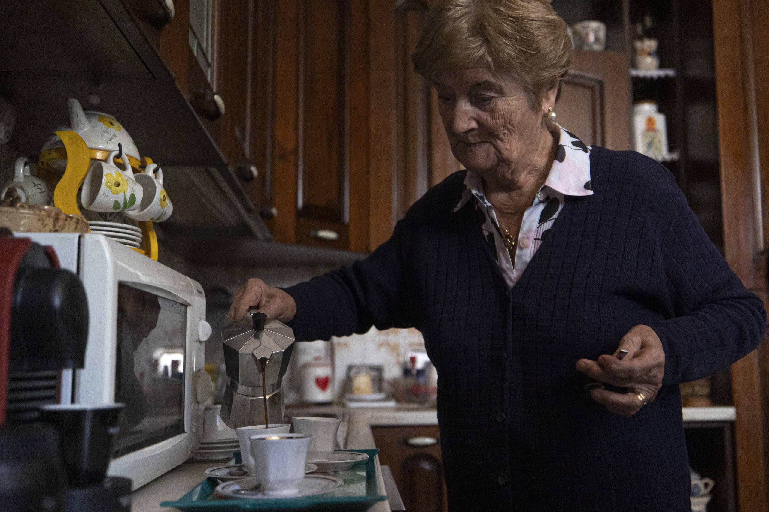 In einer gemütlichen, schwach beleuchteten Küche mit verschiedenen Küchengeräten und Dekor gießt eine ältere Frau Kaffee aus einer Espressomaschine in eine Tasse. © Fotografie Tomas Rodriguez