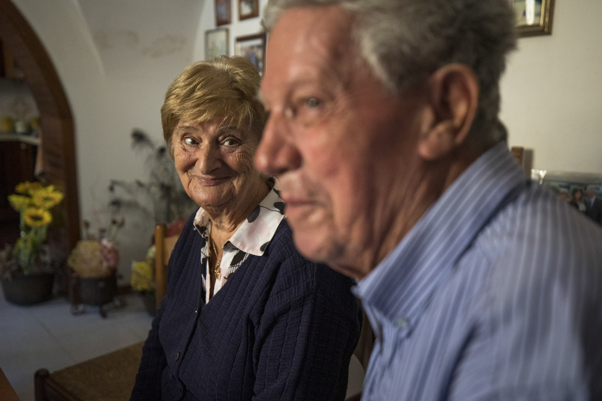 Ein älteres Ehepaar sitzt zu Hause. Die Frau lächelt herzlich in die Kamera und der Mann blickt zur Seite. Beide sind bequem gekleidet und sitzen in einer gemütlichen Umgebung. © Fotografie Tomas Rodriguez
