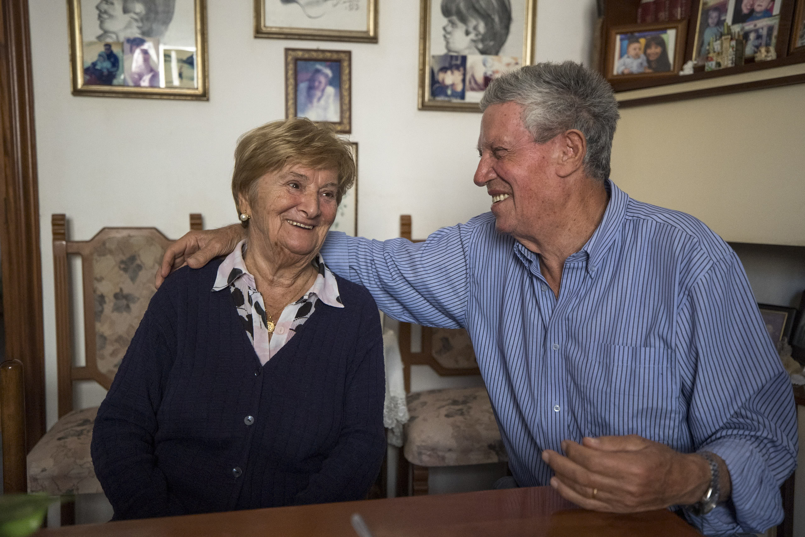 Ein älteres Paar sitzt herzlich lächelnd an einem Tisch in einem gemütlichen Raum mit Familienfotos im Hintergrund. Der Mann hat seinen Arm liebevoll um die Frau gelegt. © Fotografie Tomas Rodriguez