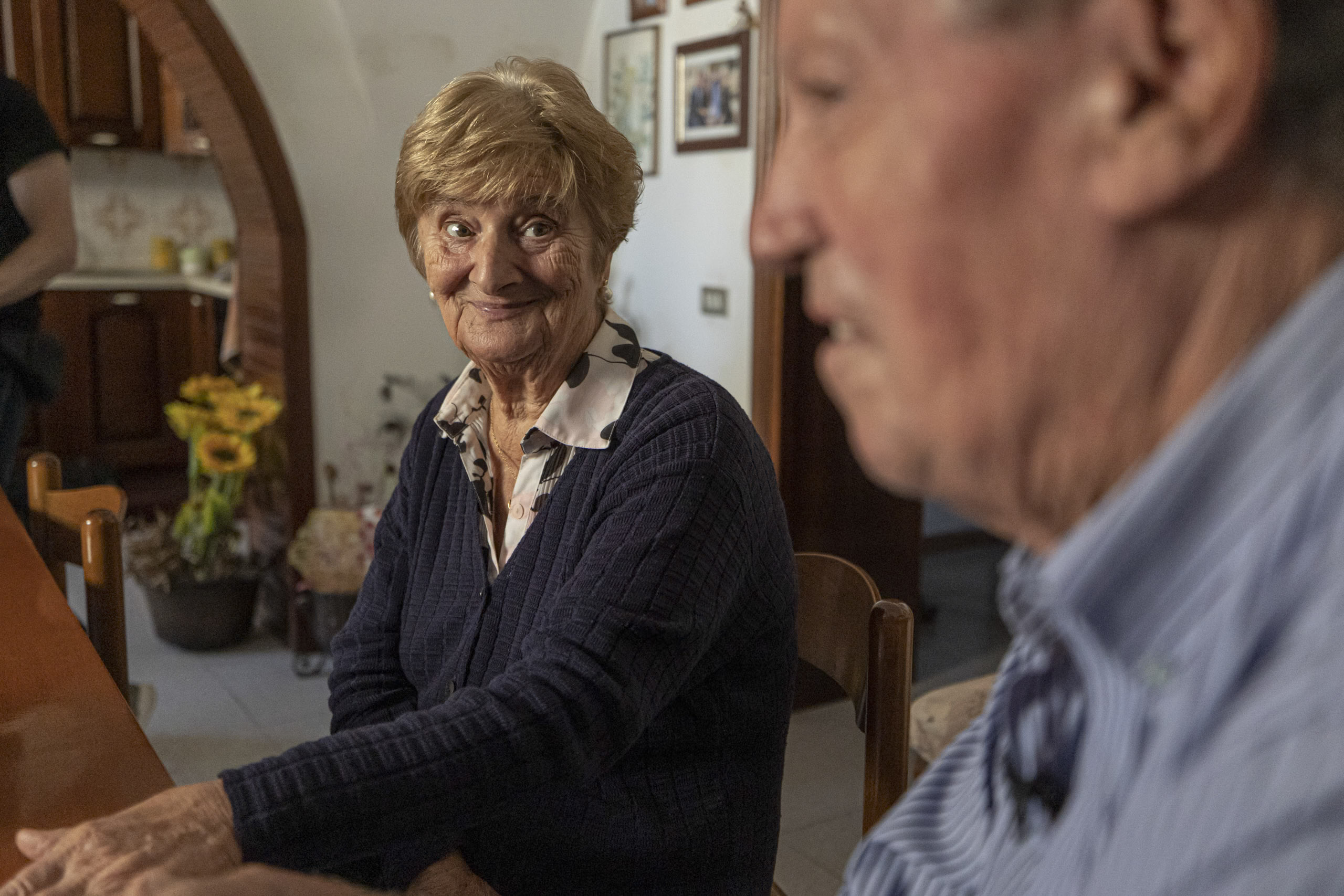 Eine ältere Frau mit kurzem, hellbraunem Haar und einem verschmitzten Lächeln sitzt an einem Esstisch und blickt in die Kamera. Ein älterer Mann, der teilweise im Vordergrund zu sehen ist, blickt von der Kamera weg. © Fotografie Tomas Rodriguez