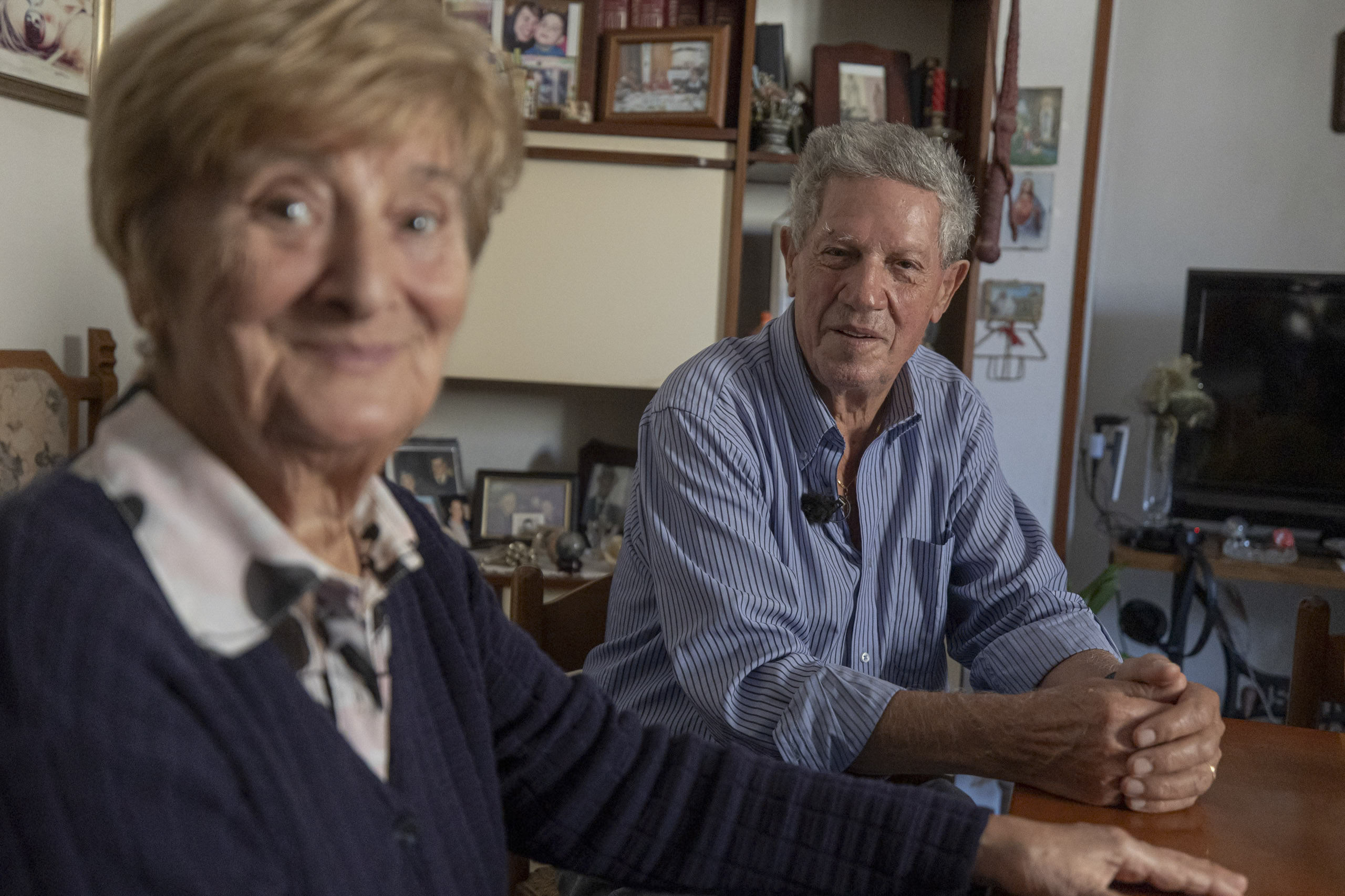Eine ältere Frau und ein älterer Mann sitzen an einem Tisch, lächeln und halten sich an den Händen, umgeben von zahlreichen gerahmten Bildern und persönlichen Gegenständen in einem gemütlichen Raum. © Fotografie Tomas Rodriguez
