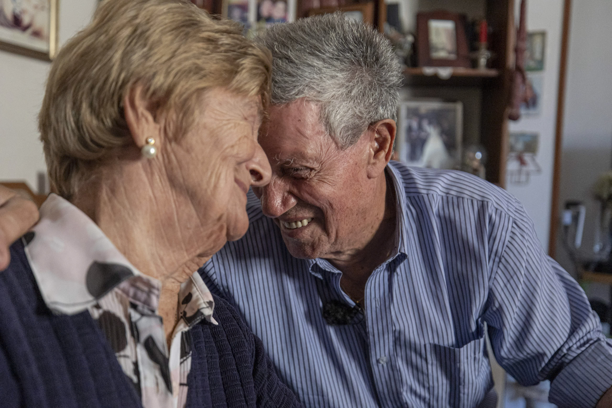 Ein älteres Paar teilt einen zärtlichen Moment, berührt die Stirn und lächelt liebevoll in einem Raum voller persönlicher Erinnerungsstücke. © Fotografie Tomas Rodriguez