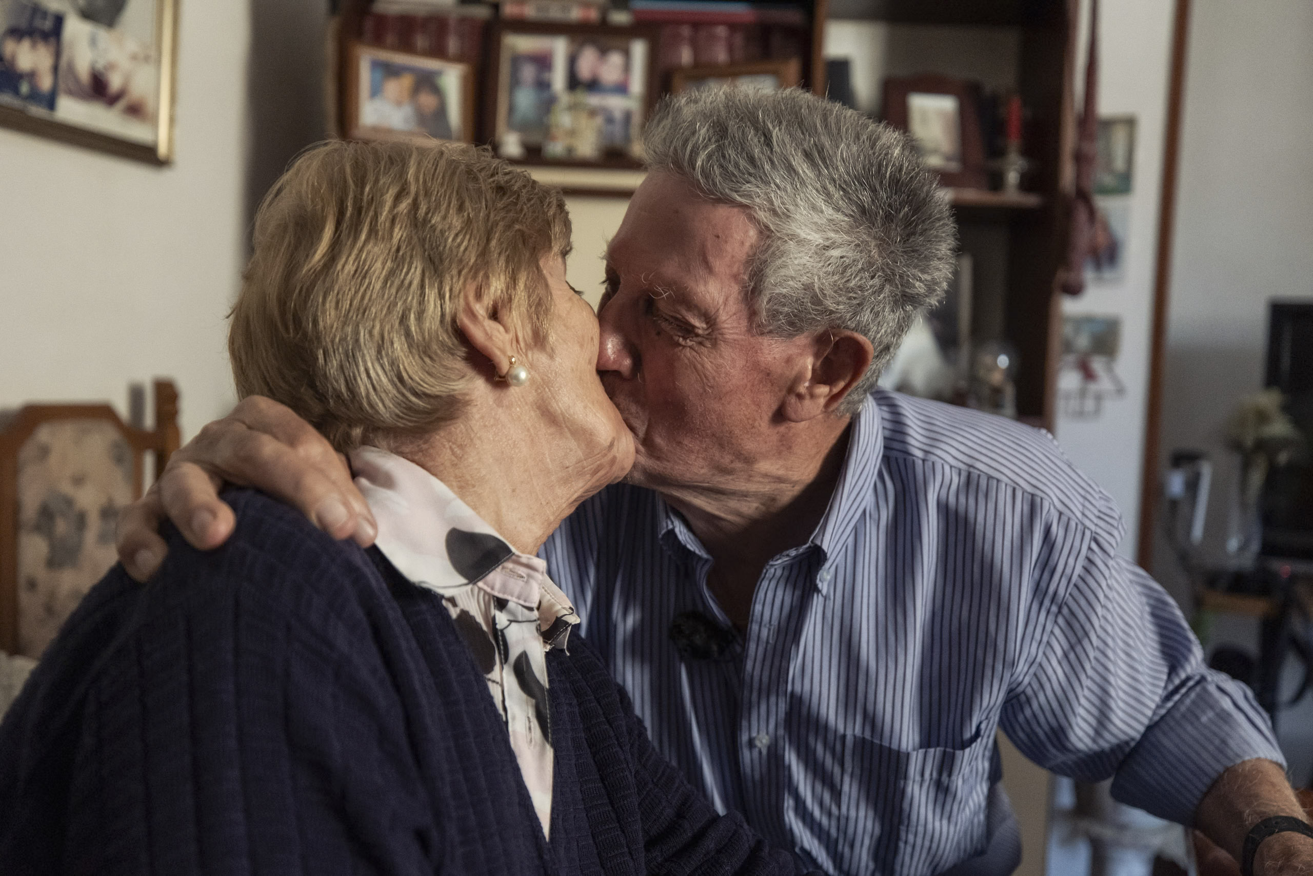 Ein älteres Paar küsst sich zärtlich in einem gemütlichen Raum voller Fotos und Dekorationen und zeigt damit einen Moment der Zuneigung und lebenslangen Kameradschaft. © Fotografie Tomas Rodriguez
