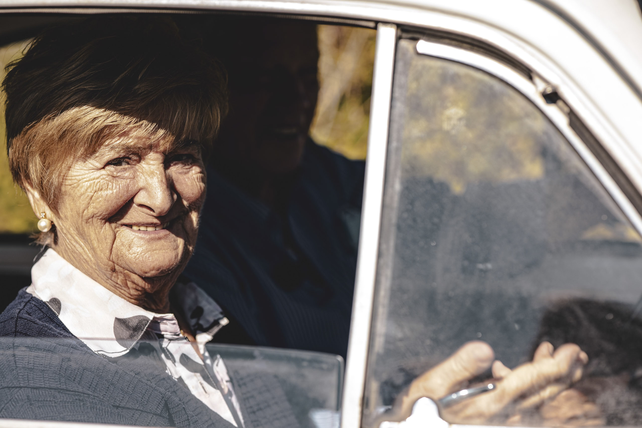 Eine ältere Frau mit fröhlichem Gesichtsausdruck sitzt auf dem Beifahrersitz eines Oldtimers. Im Hintergrund ist ihr Mann beim Fahren zu sehen. Sonnenlicht beleuchtet ihr Gesicht durch das Fenster. © Fotografie Tomas Rodriguez