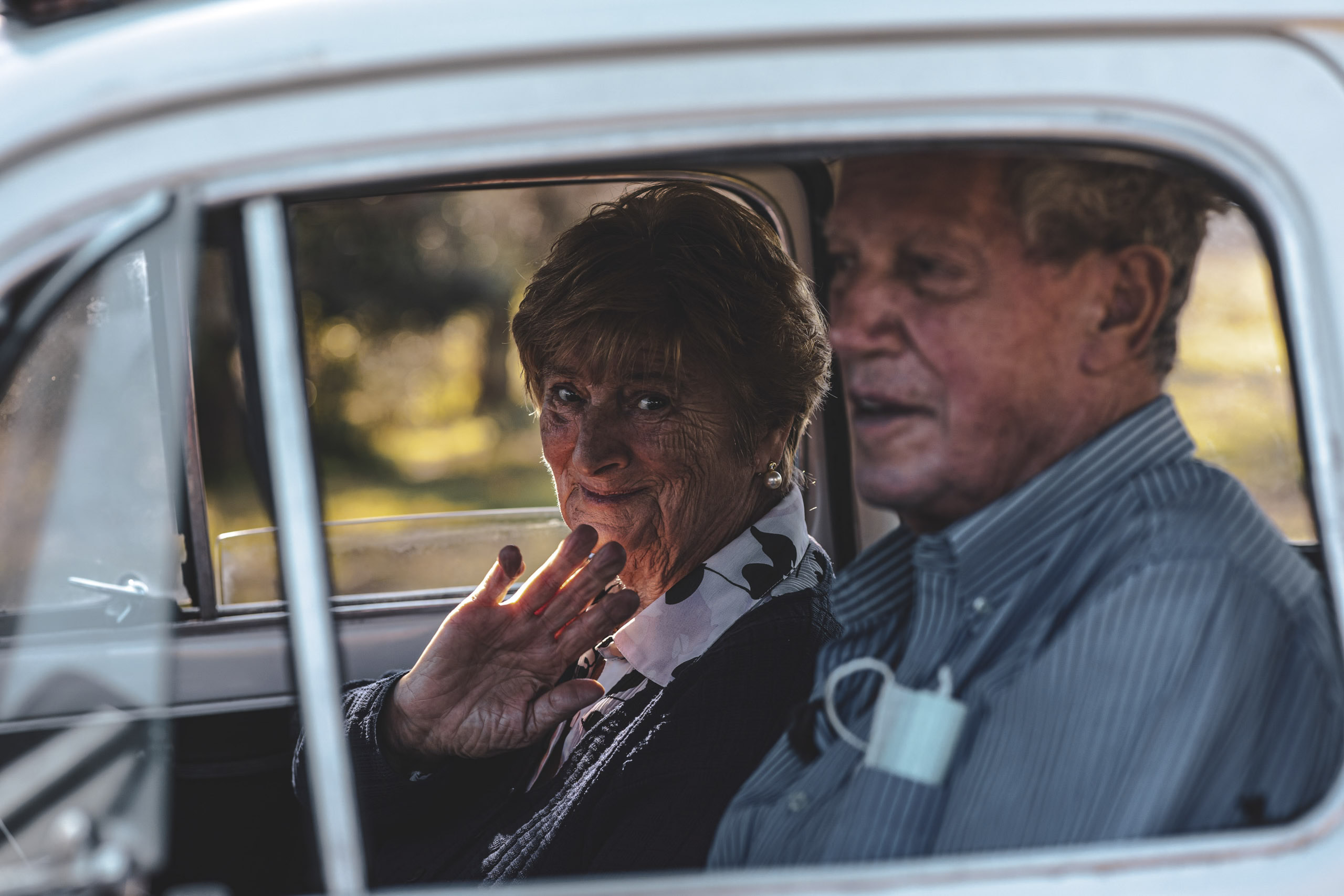 Ein älteres Ehepaar in einem Oldtimer, die Frau lächelt und schaut aus dem Fenster, der Mann sitzt am Steuer, beide in sanftes Sonnenlicht getaucht. © Fotografie Tomas Rodriguez