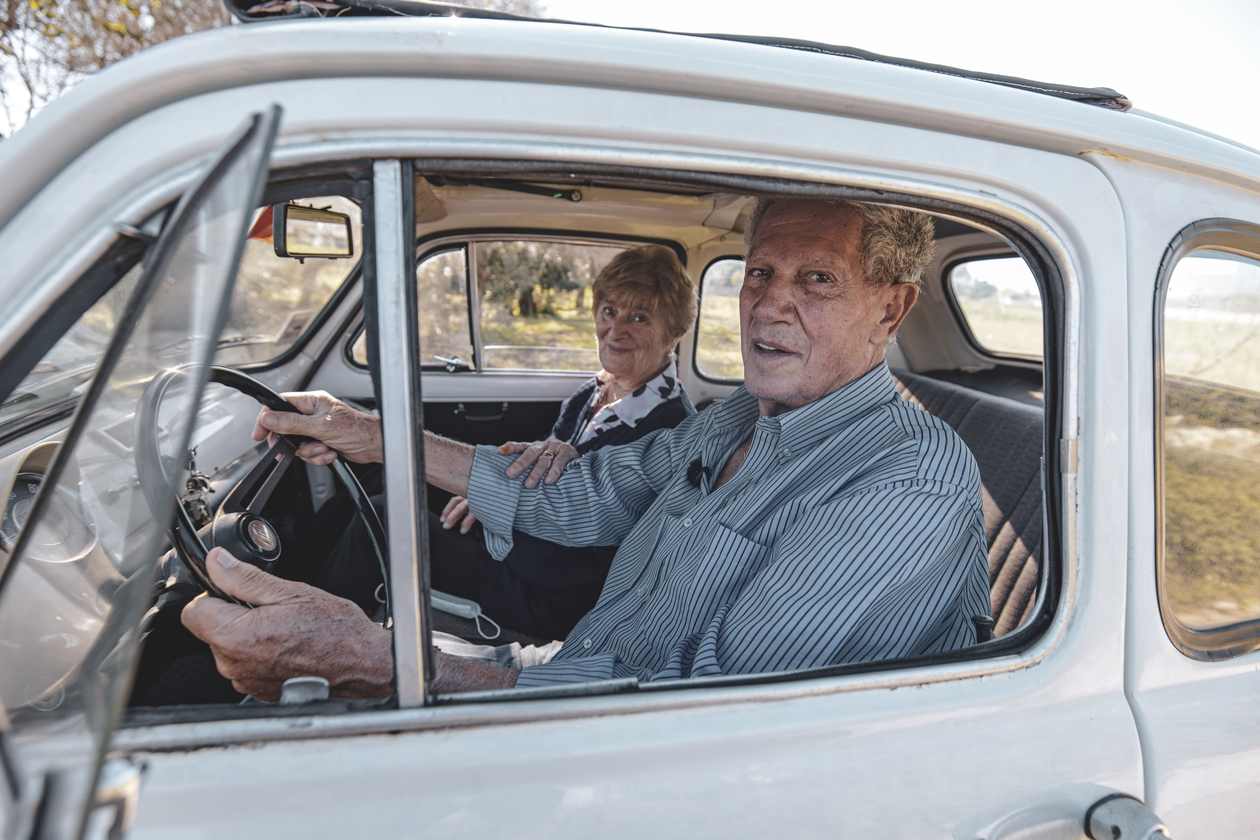 Ein älteres Paar sitzt glücklich in einem altmodischen Auto, wobei der Mann fährt und die Frau auf dem Beifahrersitz sitzt und aus dem Fenster schaut. Sie sind von einer sonnigen, natürlichen Landschaft umgeben. © Fotografie Tomas Rodriguez