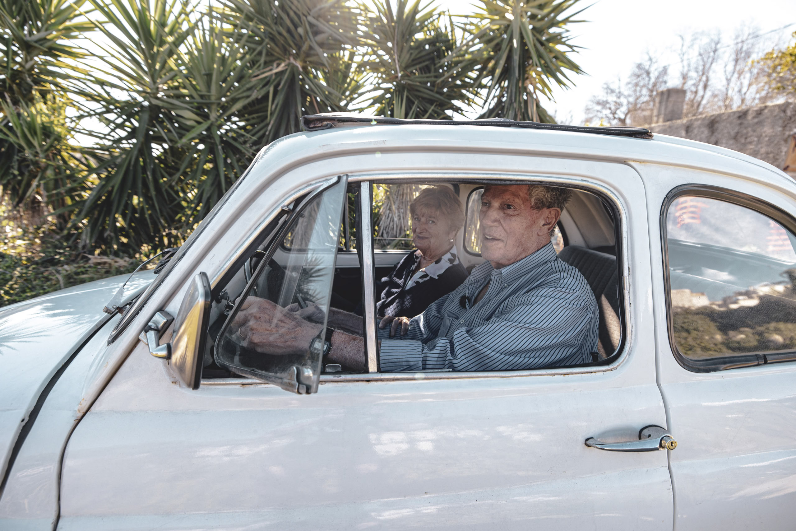 Ein älteres Paar in einem weißen Oldtimer; der Mann fährt und schaut durch das offene Fenster, die Frau sitzt auf dem Beifahrersitz, beide blicken nach vorne. © Fotografie Tomas Rodriguez