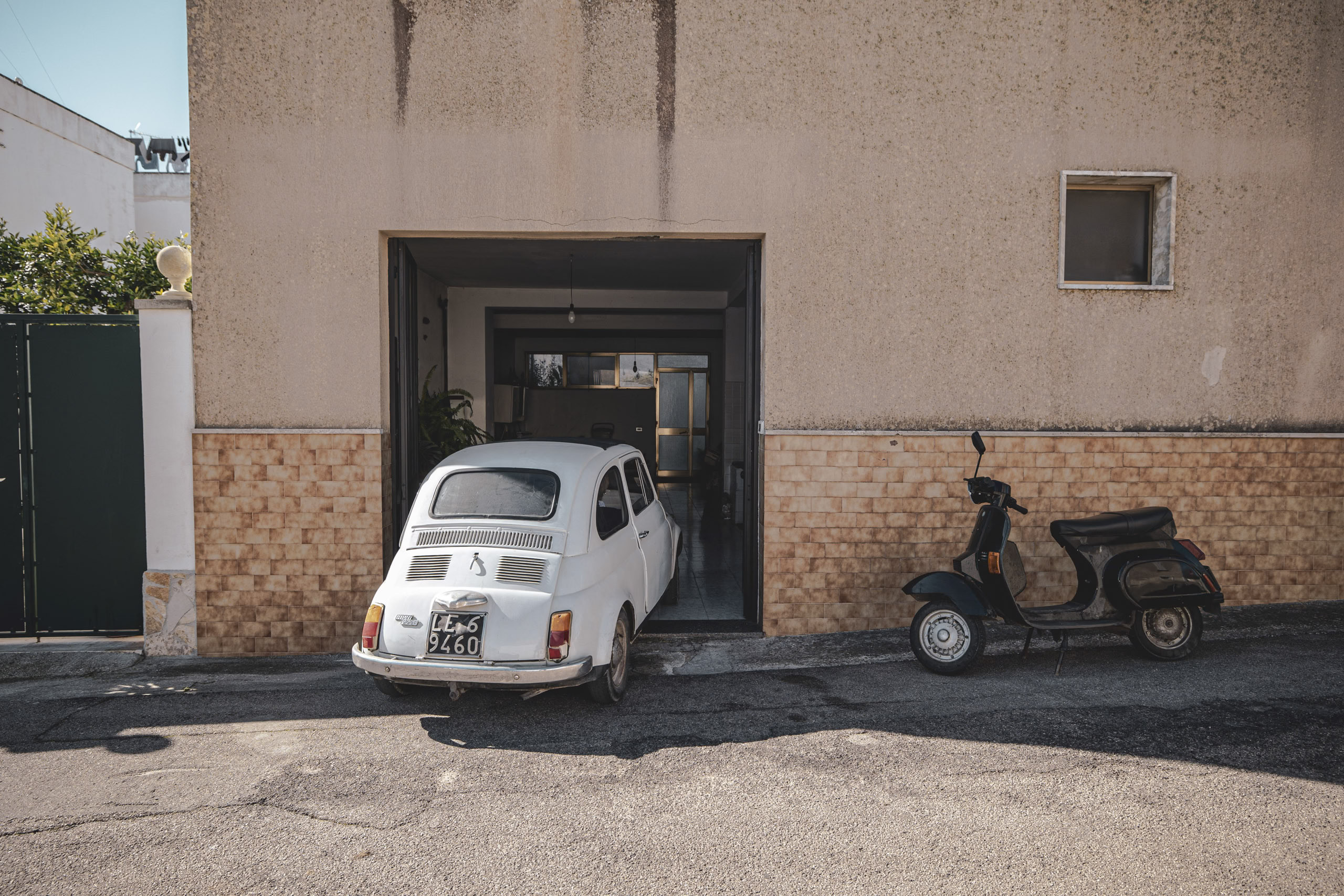 Ein weißer Oldtimer-Fiat 500 und ein Motorroller parken neben einer offenen Garage in einem sonnenbeschienenen städtischen Innenhof. Die rustikalen Gebäudewände weisen Abnutzungserscheinungen auf. © Fotografie Tomas Rodriguez