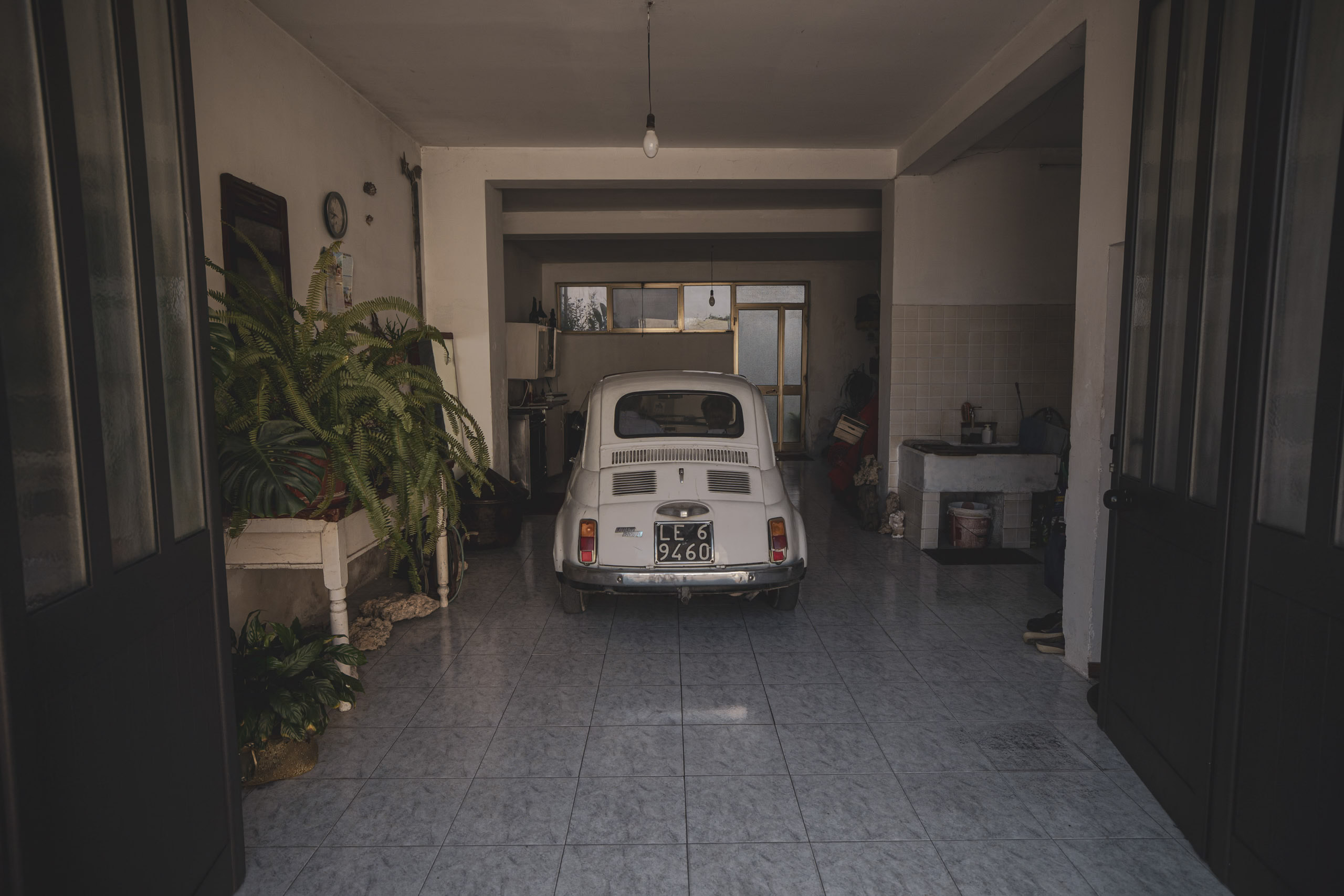 Ein weißer Oldtimer, der in einer geräumigen, gefliesten Garage geparkt ist. Die offenen Türen führen in einen sonnigen Innenhof, und links sind Zimmerpflanzen zu sehen. © Fotografie Tomas Rodriguez