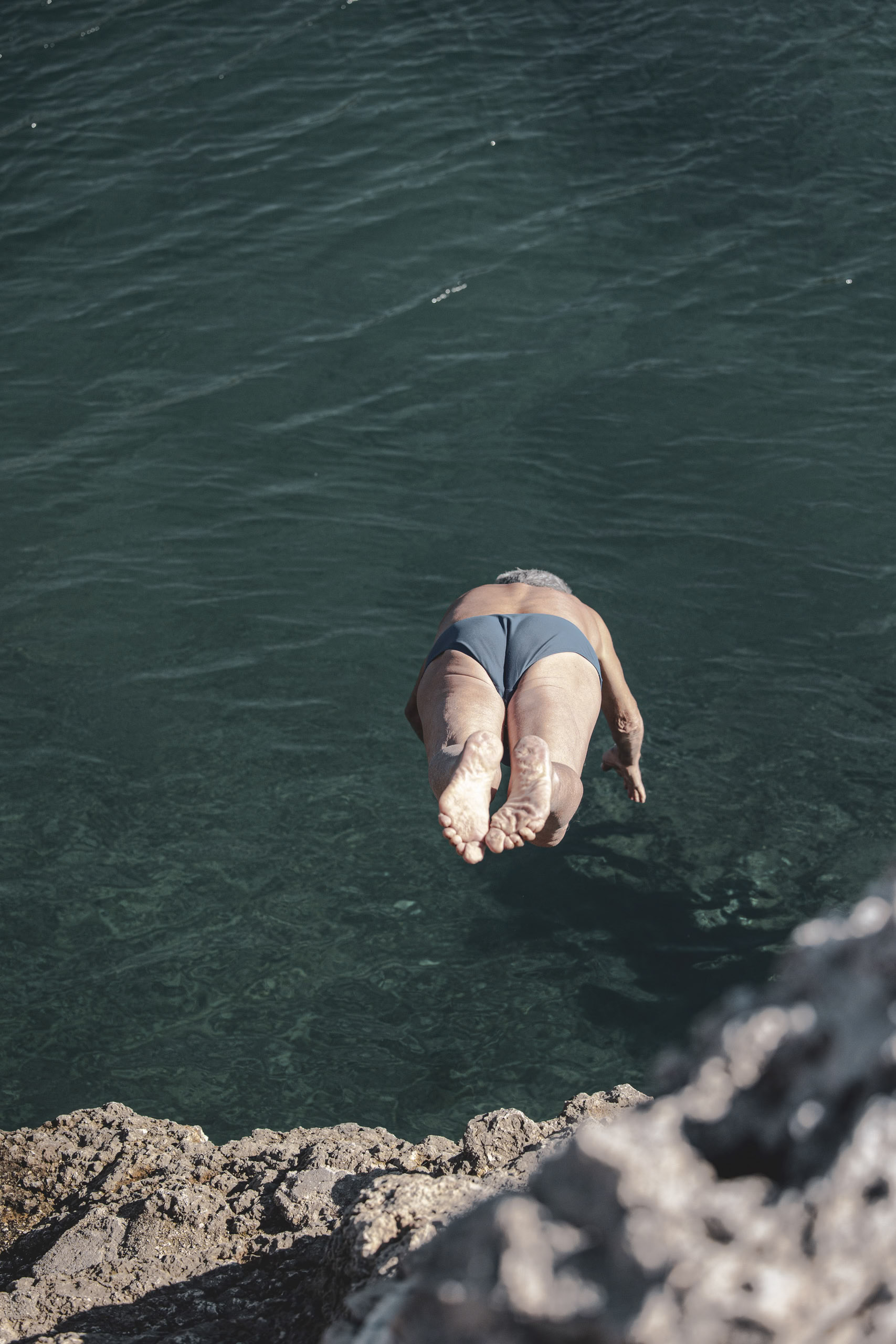Ein Mann im Badeanzug springt kopfüber von einer Felsklippe ins klare, blaue Wasser. Das Bild wurde aus einer hohen Perspektive aufgenommen, sodass man sieht, wie seine Hände zuerst ins Wasser eintauchen. © Fotografie Tomas Rodriguez